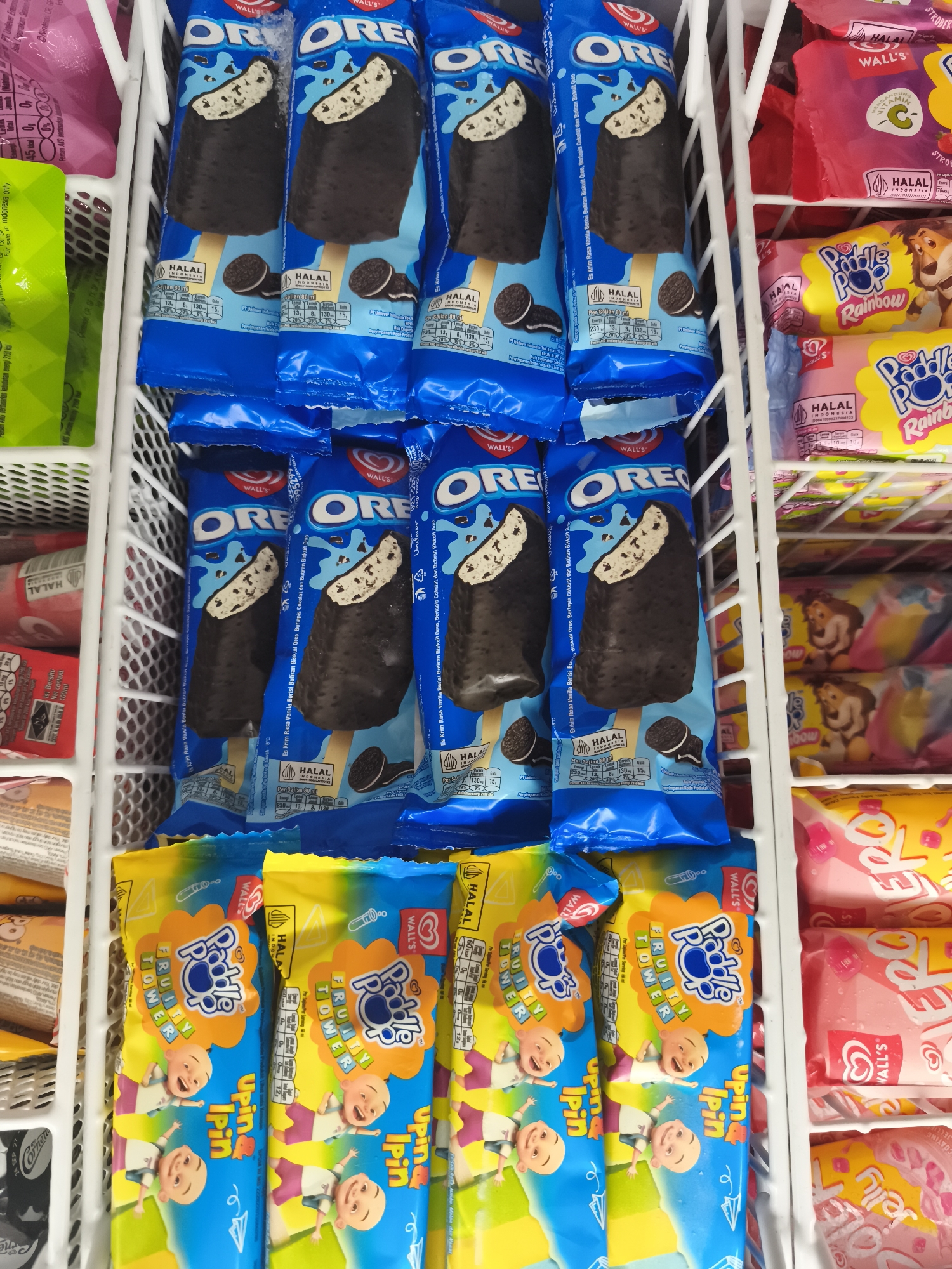 The image displays a close-up, eye-level shot of a freezer chest filled with various packaged ice cream products. The freezer is divided into sections by white wire racks. Dominating the foreground are several blue-wrapped Oreo ice cream bars, each showing a portion of the ice cream with chocolate coating and visible cookie pieces. Below these, brightly colored yellow and blue packages of "Paddle Pop Fruity Tower" ice cream, featuring cartoon characters, are visible. To the right, pink and yellow packaged ice cream bars with text like "WALL'S" and "Jelly" are stacked. The background shows more ice cream packaging, including some pink items at the top left. The scene appears to be set in a retail environment, likely a convenience store or supermarket. The specific location is identified as Kemiri Kidul, Indonesia, suggesting a local Indonesian market setting. There are no people or overt actions depicted, the focus is solely on the assortment of frozen treats.