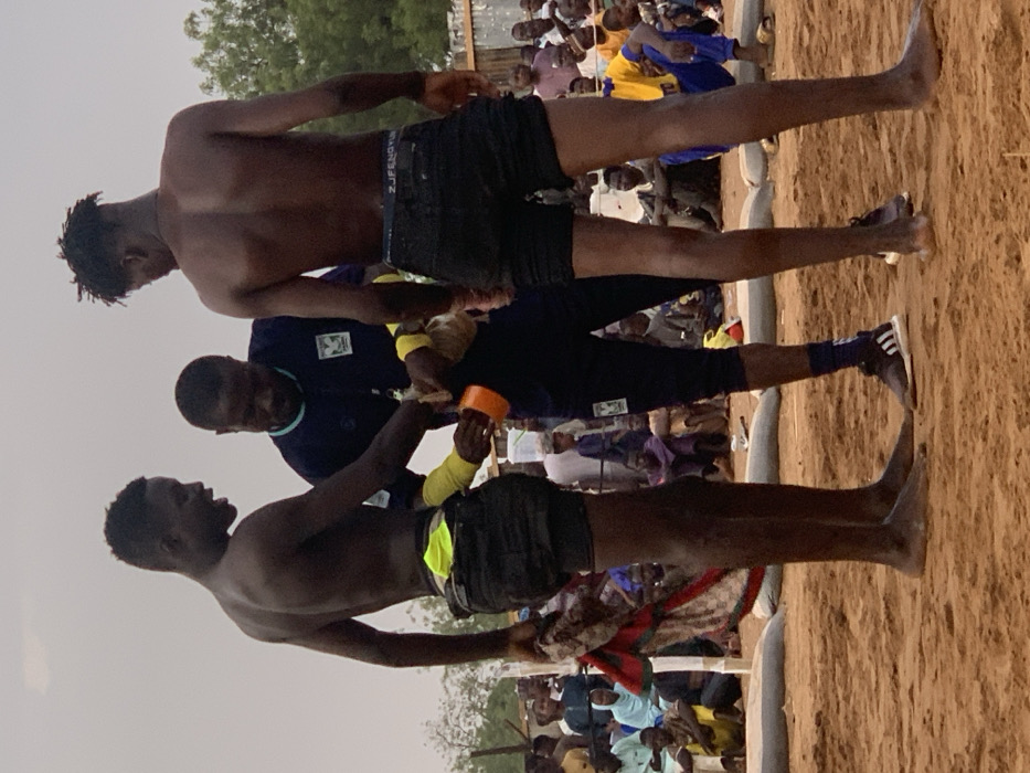 The image shows three men standing on a sandy wrestling ring. The man on the left is shirtless and wearing black shorts with a yellow waistband, holding a red and white cloth in his left hand. He is looking down at the man in the middle. The man in the middle is wearing a dark blue tracksuit with a white logo. He is wrapping something around the man on the right's right wrist. The man on the right is shirtless and wearing black shorts. His back is to the camera. The man in the middle is holding an orange object in his left hand. The three men are standing on a sandy wrestling ring. There is a crowd of people sitting in the background. The image appears to have been taken in a rural area. The coordinates 13.049, 5.237 place the image in Sokoto, Nigeria.