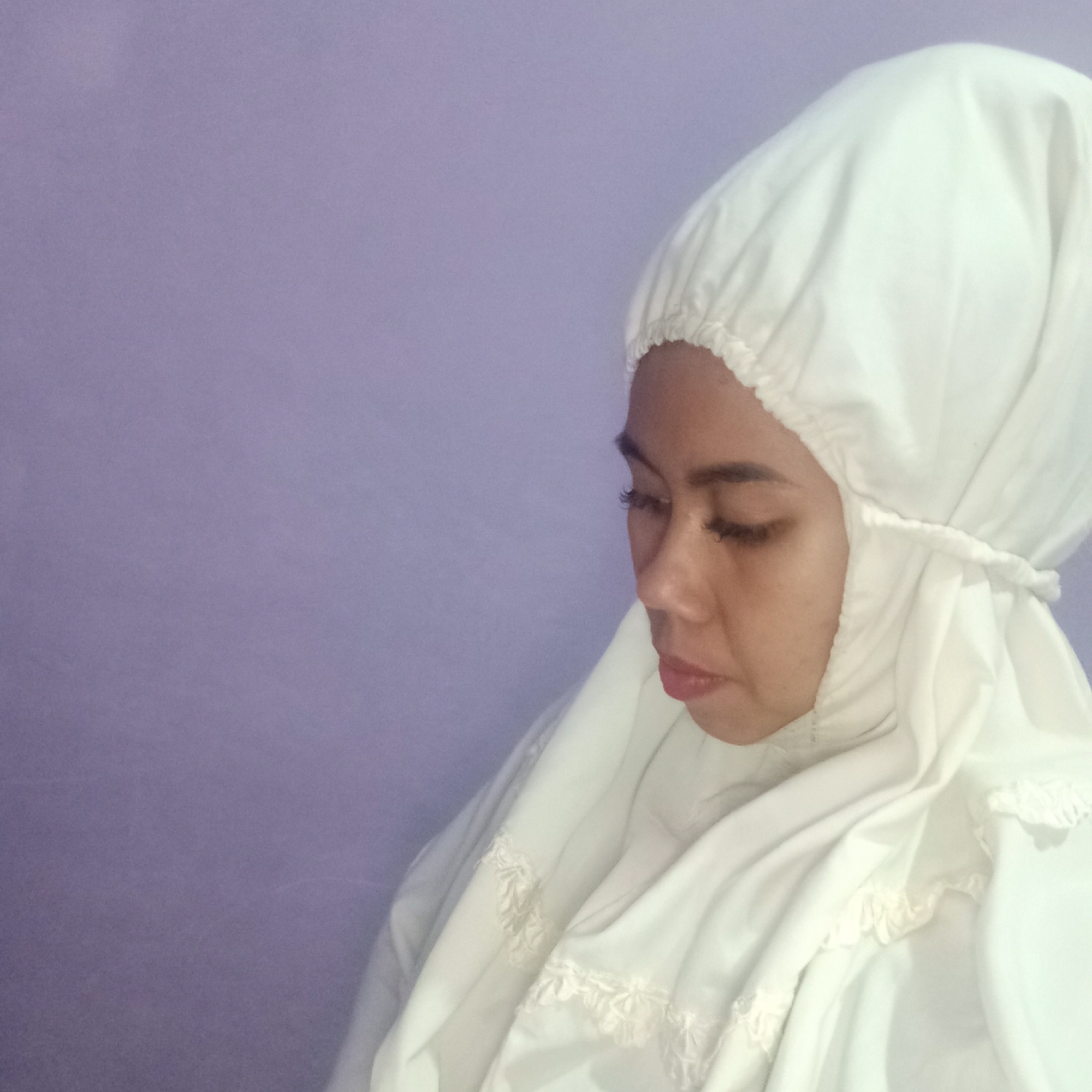 A young Indonesian woman of East Asian descent is captured in a medium shot, her head tilted downwards and her eyes closed, wearing a white mukena (prayer garment). The mukena covers her head and neck, featuring a white lace trim along the hem and a drawstring detail around the head covering. Her skin tone is light brown, and she has dark, arched eyebrows and long eyelashes. The background is a plain, solid purple wall, suggesting an indoor setting. The lighting is soft, casting gentle shadows and highlighting the texture of the fabric. The image appears to be taken in Tegowanu Kulon, Indonesia, likely in a residential setting.