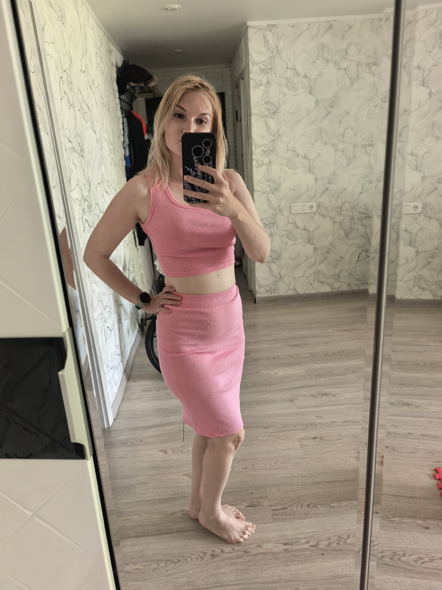 A blonde woman stands barefoot in a room, posing in front of a mirror for a photo taken with a smartphone. She wears a pink, two-piece outfit consisting of a tank top and a skirt. The room features a light-colored wooden floor, walls with a marble-like pattern, and a wardrobe with mirrored doors on the left. In the background, a clothes rack and a glimpse of an adjacent room are visible. The location is likely an apartment or home in Sterlitamak, Russia.