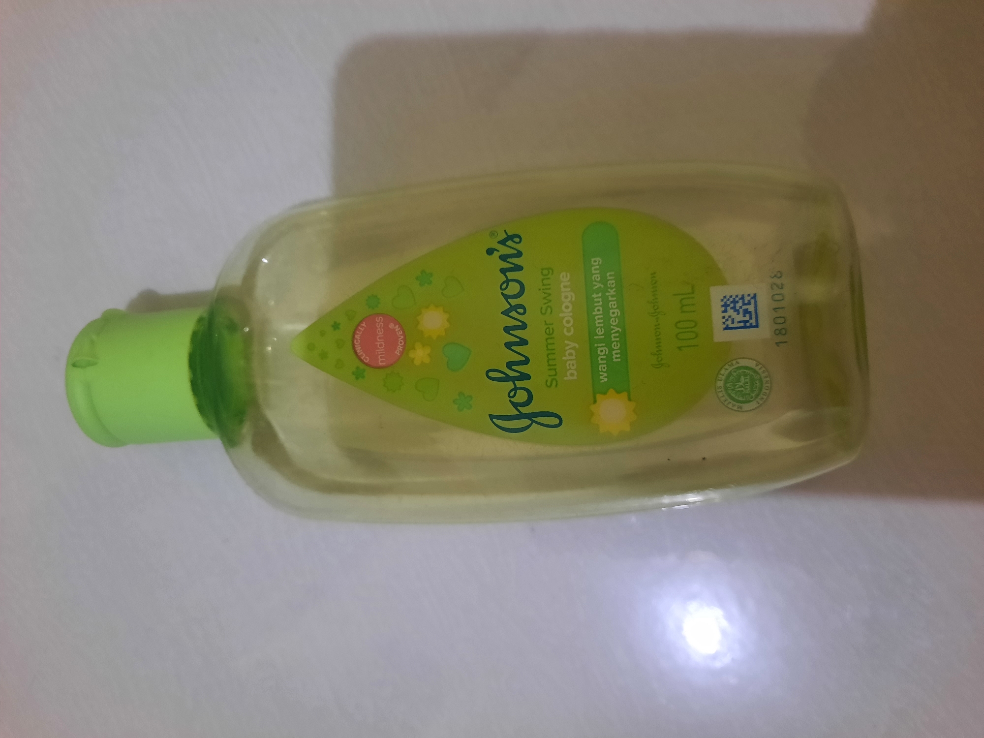 A Johnson's Baby Cologne bottle, 100 mL, with a green cap and label, is shown from a slightly elevated angle. The label features the "Johnson's" logo, "Summer Swing baby cologne," and Indonesian text that translates to "mild fragrance that refreshes." A halal certification logo and a QR code are also visible on the bottle. The background is a white, textured surface, possibly a tabletop. There are no people or activities depicted in the image. The bottle is positioned on the surface, with a bright reflection suggesting artificial lighting. The overall scene is a product shot of the baby cologne. The location within the city of Lhokseumawe, Indonesia, is not discernible from the provided image.