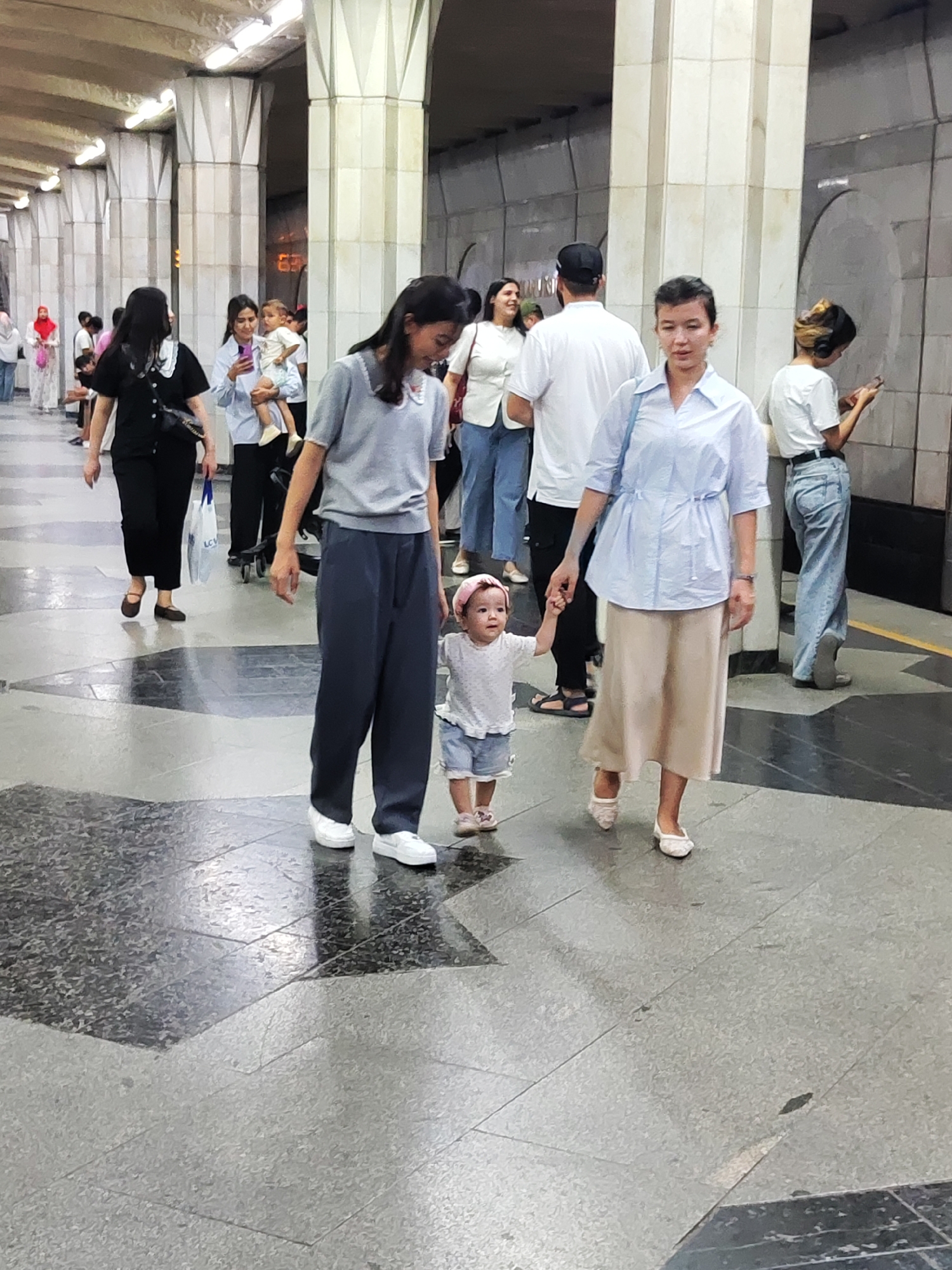 The image captures a street scene in Tashkent, Uzbekistan, likely within a metro station or underpass, characterized by its distinctive tiled flooring and large architectural columns. In the foreground, a woman in a light blue collared shirt and a beige skirt is holding the hand of a young child wearing a pink headband and a white t-shirt with denim shorts. They are walking on a polished floor with a patterned design. Behind them, another woman in a grey top and dark trousers is walking, and further back, several other individuals are visible, including a woman in a black outfit and a woman holding a baby. The environment suggests an urban public space, with a sense of movement and everyday life.