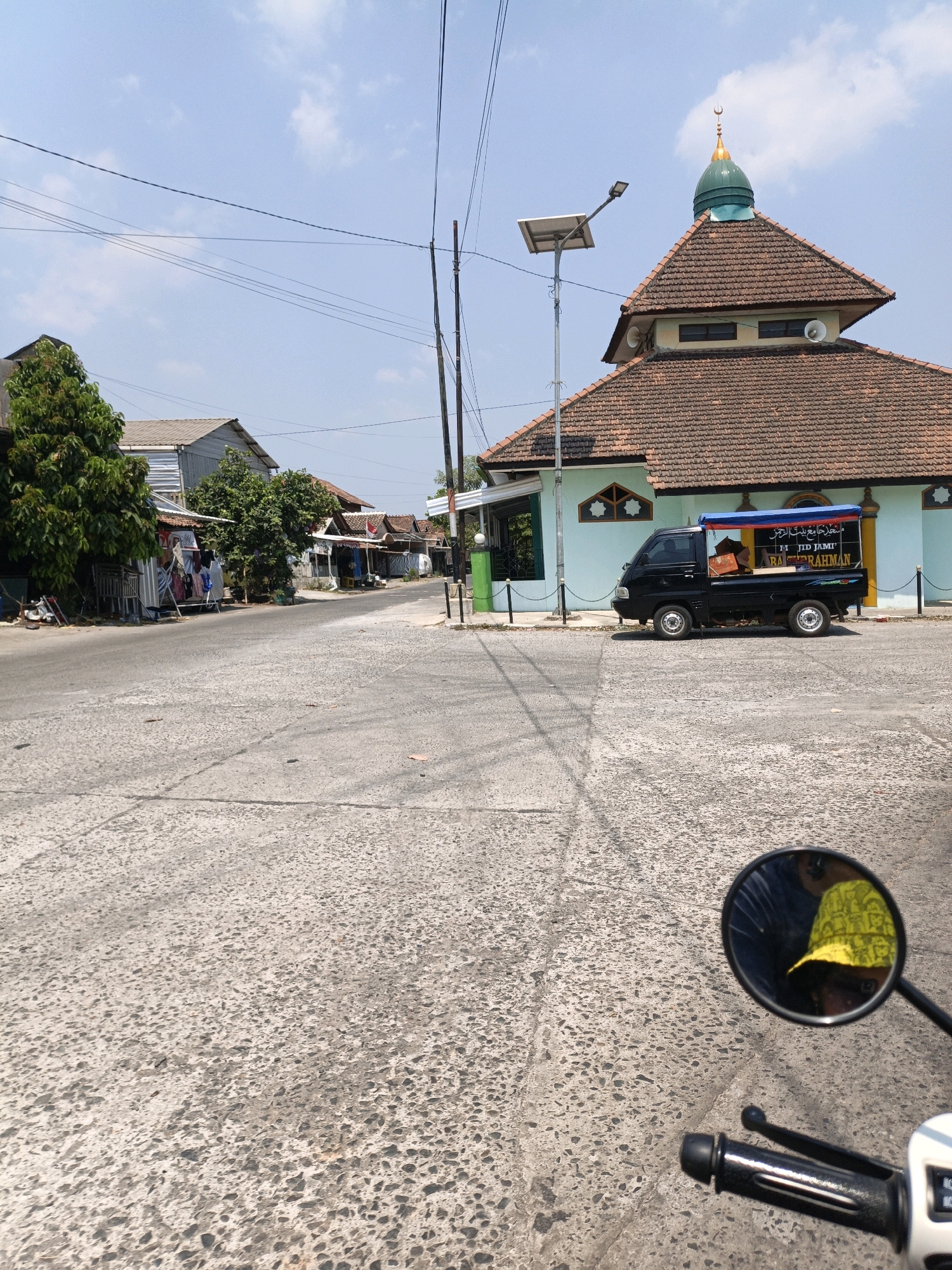 This is a street-level, eye-level shot taken from a motorcycle in Jepara, Indonesia. The image captures a mosque with a green dome and brown tiled roof on the right side of the frame. A black pickup truck is parked in front of the mosque. The street is paved with concrete and shows signs of wear. On the left side of the street, there are several small shops and trees. The sky is clear with a few scattered clouds. The reflection of a person wearing a yellow hat can be seen in the motorcycle's rearview mirror in the lower right corner.