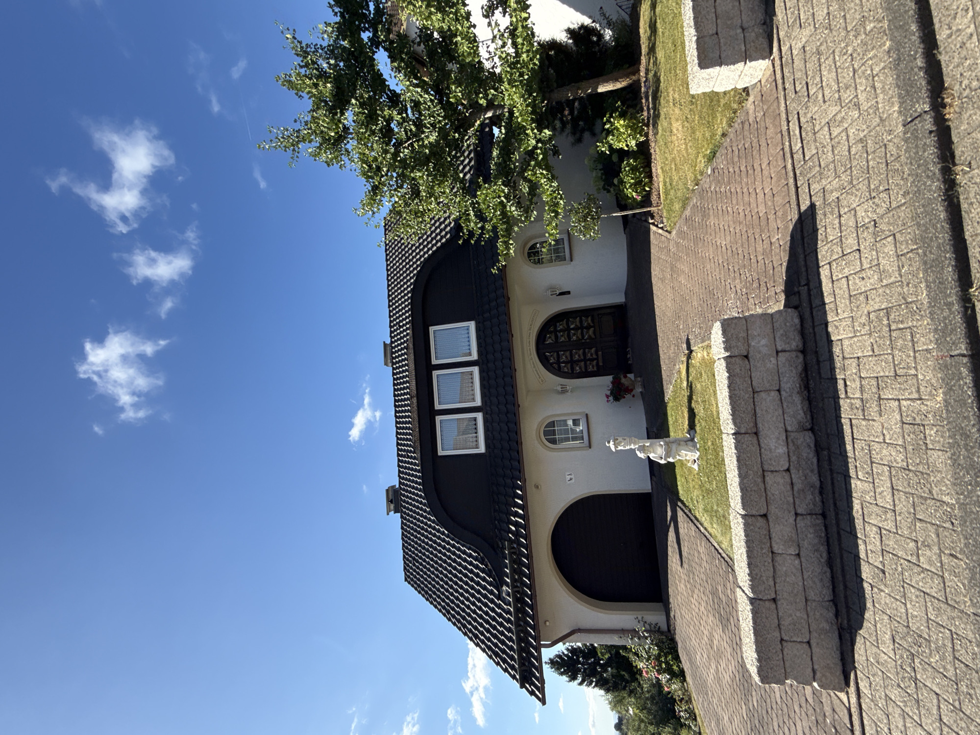 A suburban detached house in Bad Sachsa, Germany, is depicted on a sunny day with a clear blue sky and scattered white clouds. The house features a dark, mansard-style roof with multiple dormer windows and a dark, arched garage door on the left. The facade is white with arched windows and a prominent, dark double door entrance. A mature tree with lush green foliage stands to the right of the house. In front of the house, a paved driveway leads to the garage and entrance. A small white statue of a child holding a basket is situated on a patch of grass between the driveway and the house. A low, grey stone retaining wall borders the driveway in the foreground. The surrounding area appears to be a residential neighborhood with greenery.