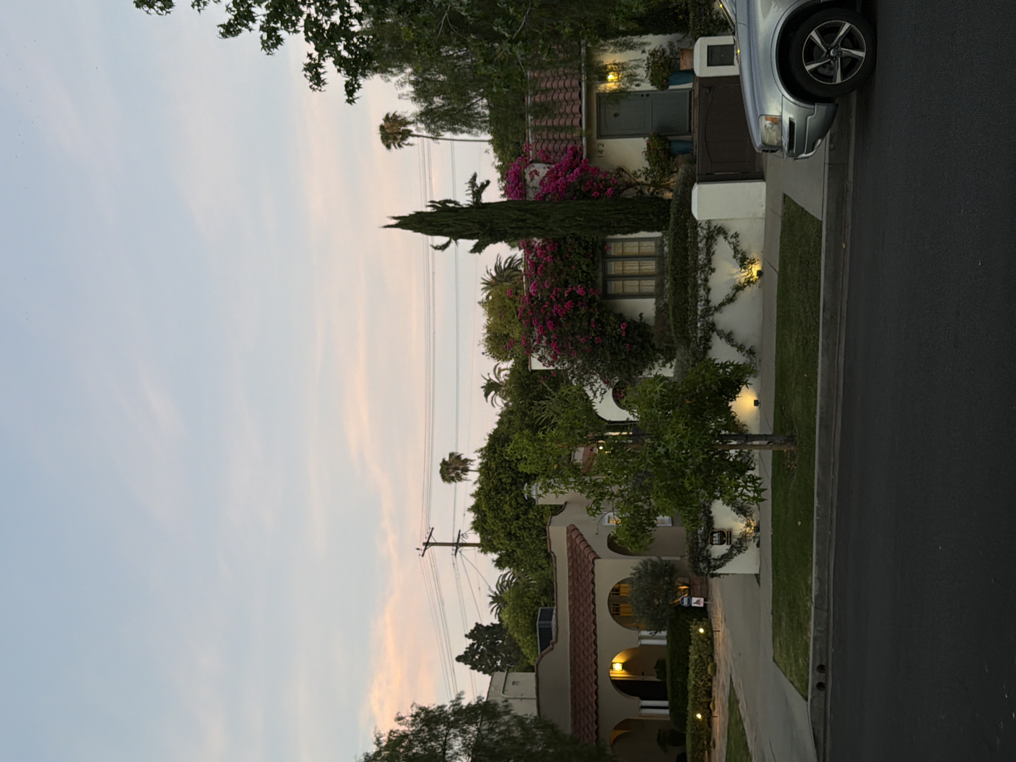 An exterior, medium shot captures a quiet residential street in the Los Angeles area, USA, at dusk. The sky is a soft gradient of pale blue to pink, with wispy clouds. On the right side of the frame, a silver SUV is parked on the street, partially visible. The house in the background, displaying the house number "142," is a single-story dwelling with white stucco walls, a red tile roof, and a dark green door. It is adorned with vibrant pink bougainvillea and a tall, slender cypress tree. The house on the left features arched entrances and a terracotta tile roof. Lush green trees and manicured hedges line the properties, with a street light visible in the foreground. A wooden utility pole with electrical wires stands in the distance. The scene is peaceful, illuminated by the soft glow of streetlights and the fading daylight.