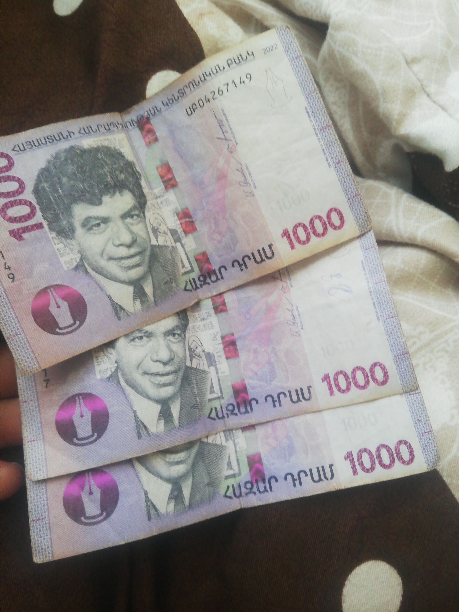 The image shows a close-up view of several Armenian dram banknotes, specifically the 1000 dram denomination. The banknotes are stacked and partially overlap each other. Each banknote features a portrait of a man, a watermark, and the denomination "1000" printed prominently. The banknotes appear to be in good condition. The background consists of a textured fabric, possibly a blanket or upholstery, with a pattern of light and dark circular shapes. The lighting suggests an indoor setting, but no specific location details are visible.