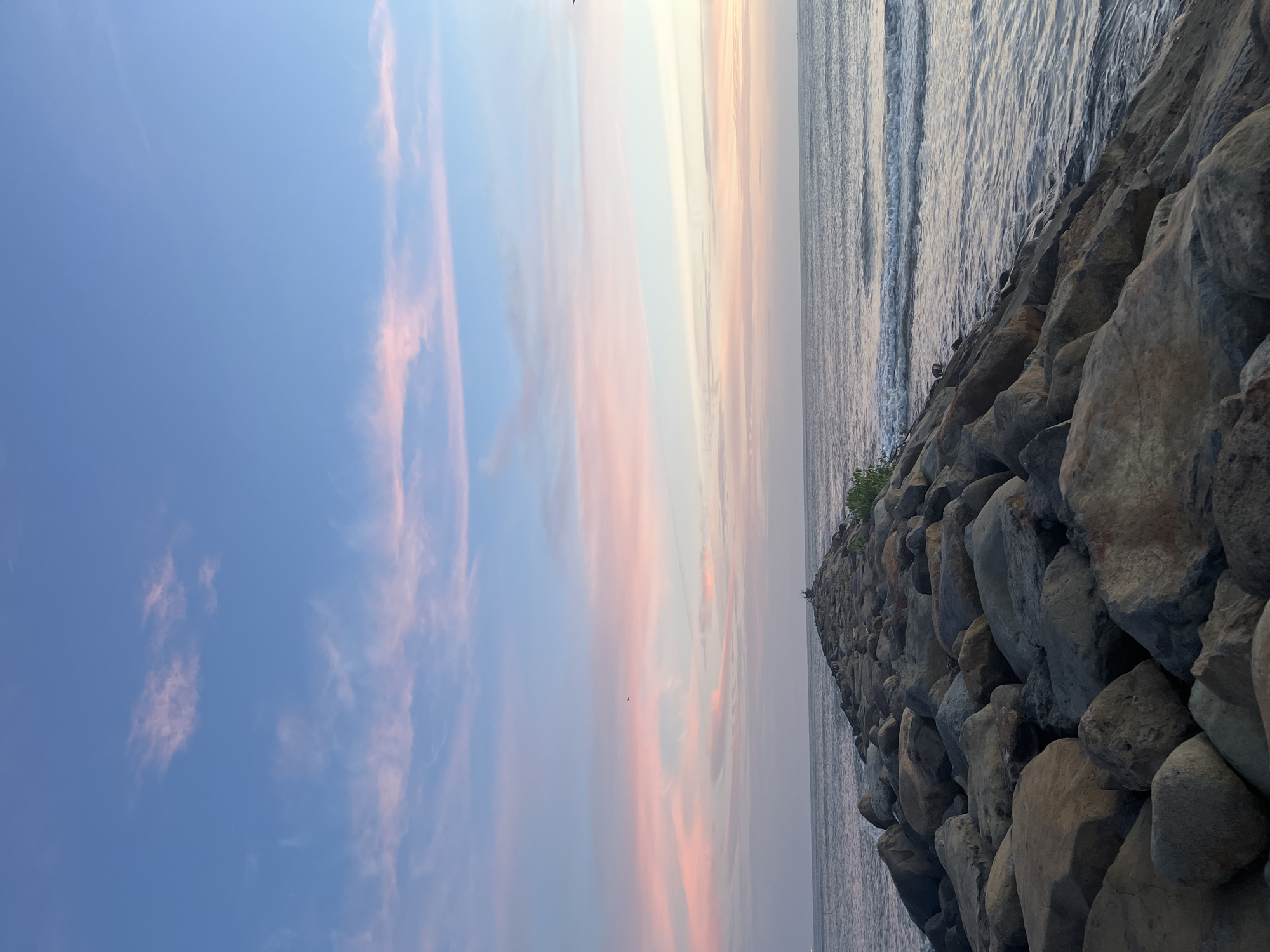 A stone jetty extends into the ocean at Keudeteupinpunti, Indonesia. The sky is a gradient of blue, pink, and white, with wispy clouds. The ocean is calm, with small waves lapping at the shore. The jetty is made of large, rough-hewn stones, some of which are covered in moss. There are a few small plants growing on the jetty. A single bird can be seen flying in the distance. The overall scene is peaceful and serene.