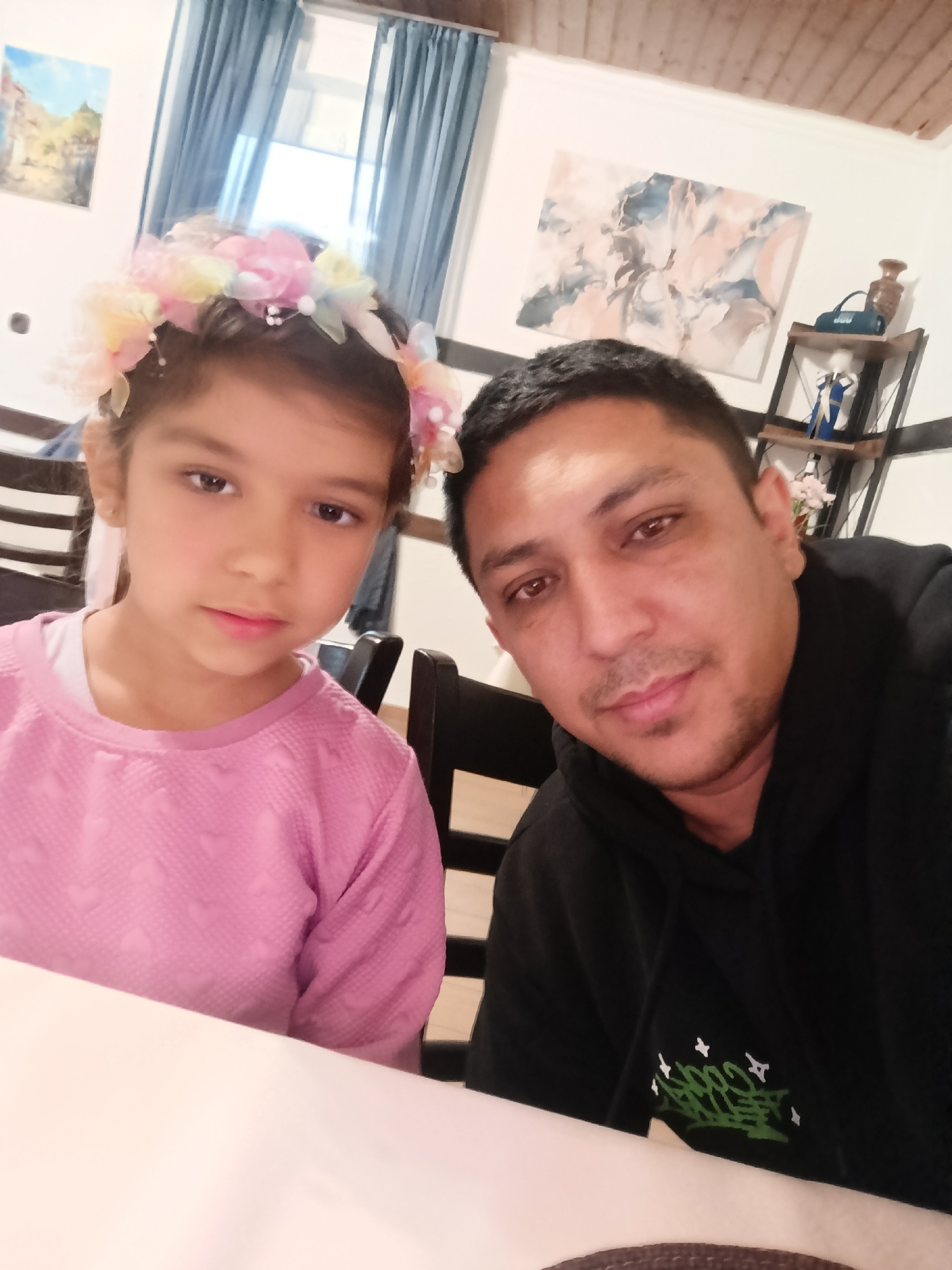 A close-up, selfie-style image shows a man and a young girl posing for a photo indoors. The girl, on the left, has dark hair and is wearing a pink, textured long-sleeved shirt. A colorful flower crown adorns her head. The man, on the right, has dark hair and is wearing a black hooded sweatshirt. Both subjects are looking towards the camera. The background suggests a casual indoor setting, possibly a dining area, with dark chairs visible and a table in the foreground. In the background, there's a painting on the wall and shelving with decorative items. The lighting is indoor, and no specific time of day or weather conditions are discernible. There are no visible landmarks to pinpoint the location within Reutlingen, Germany. The mood appears relaxed and familial. A small green graphic design is visible on the man's sweatshirt.