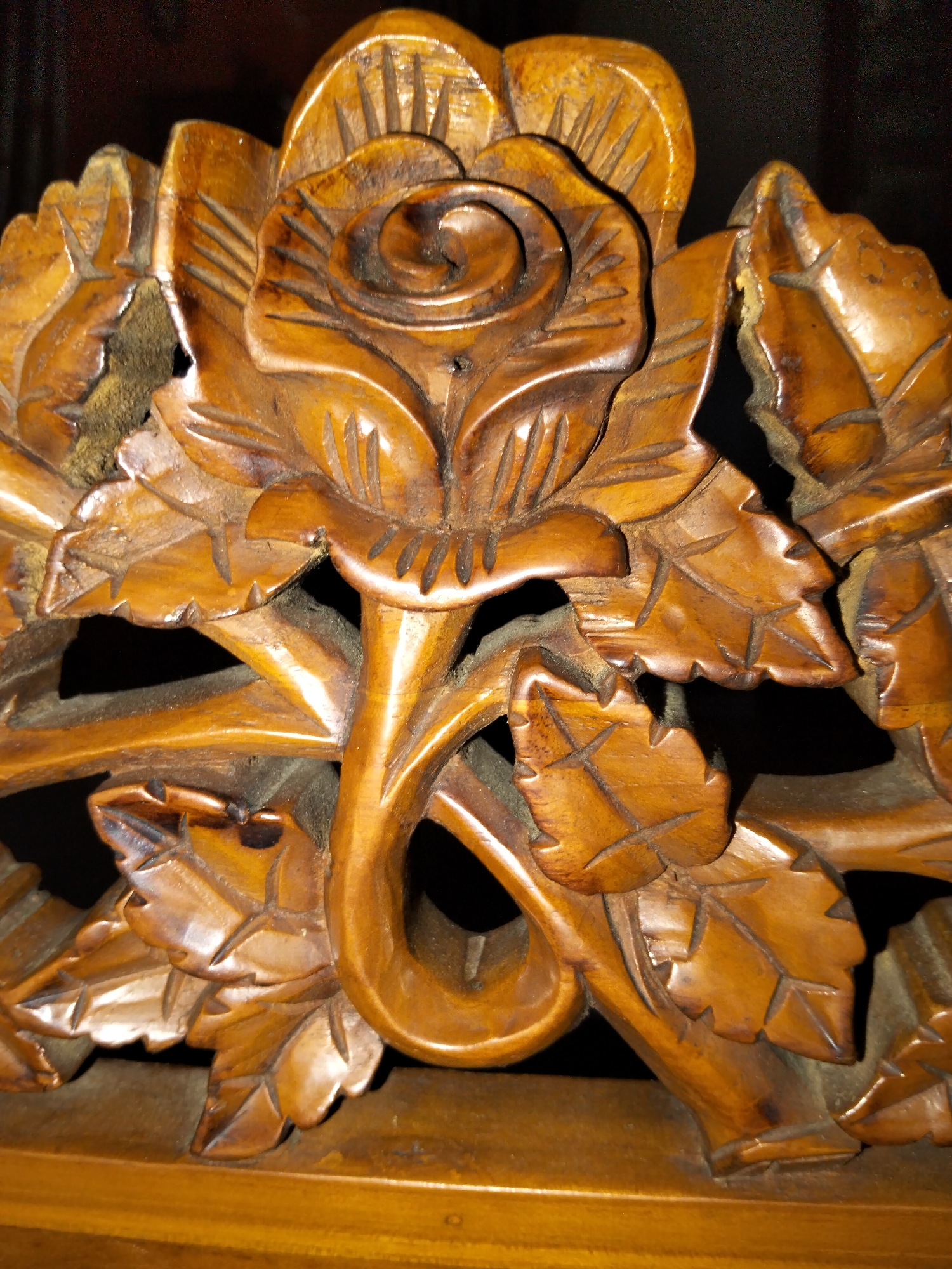 The image displays a close-up, detailed view of intricate wood carving, likely an ornamental piece. The central subject is a stylized rose with layered petals, surrounded by carved leaves and stems. The wood appears to be polished and has a warm, reddish-brown hue, with visible grain patterns and some darker patina in the recessed areas, suggesting age or intentional staining. The carving shows signs of meticulous craftsmanship, with sharp lines defining the edges of the petals and veins of the leaves. Some areas appear to have a fuzzy, greenish-brown material lodged within crevices, possibly moss or aged dust accumulated over time, particularly noticeable on the left side. The lighting casts deep shadows, highlighting the three-dimensional texture and depth of the carving. The background is dark and indistinct, further emphasizing the carved object. The object is resting on a polished wooden surface, which reflects some of the light. No people or specific geographical features of Payagaboh, Indonesia, are visible in this close-up, detailed shot of the carved artifact.