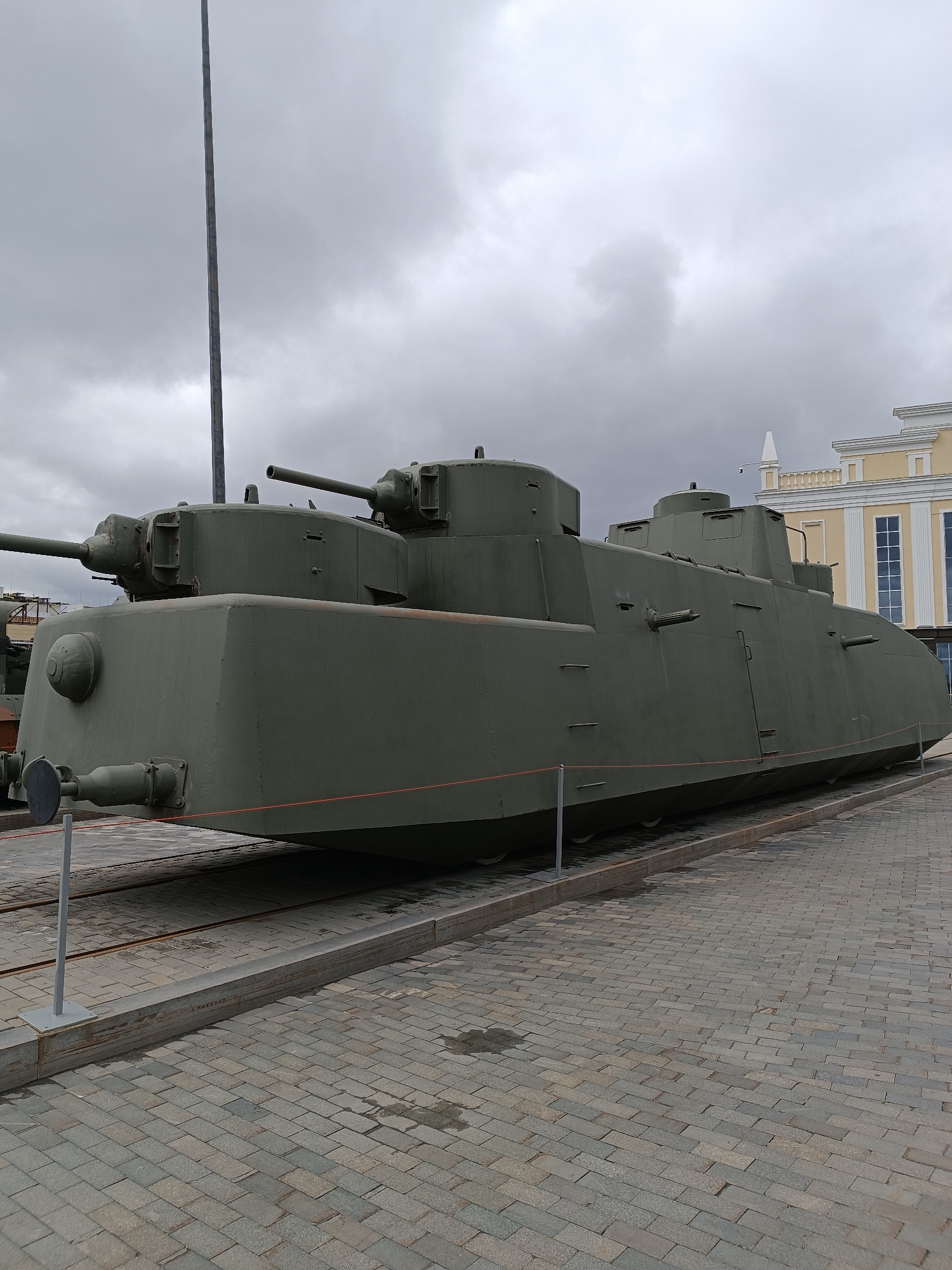 The image shows a large, olive-drab armored train on display in Verkhnyaya Pyshma, Russia. The train is positioned on railway tracks, with a paved walkway in the foreground and a building in the background. The armored train appears to be a museum exhibit, with various turrets and gun emplacements visible along its length. The sky is overcast, contributing to a somewhat muted color palette. The building in the background is light-colored with a classical architectural style, featuring arched windows and decorative elements. There are no people visible in the image.