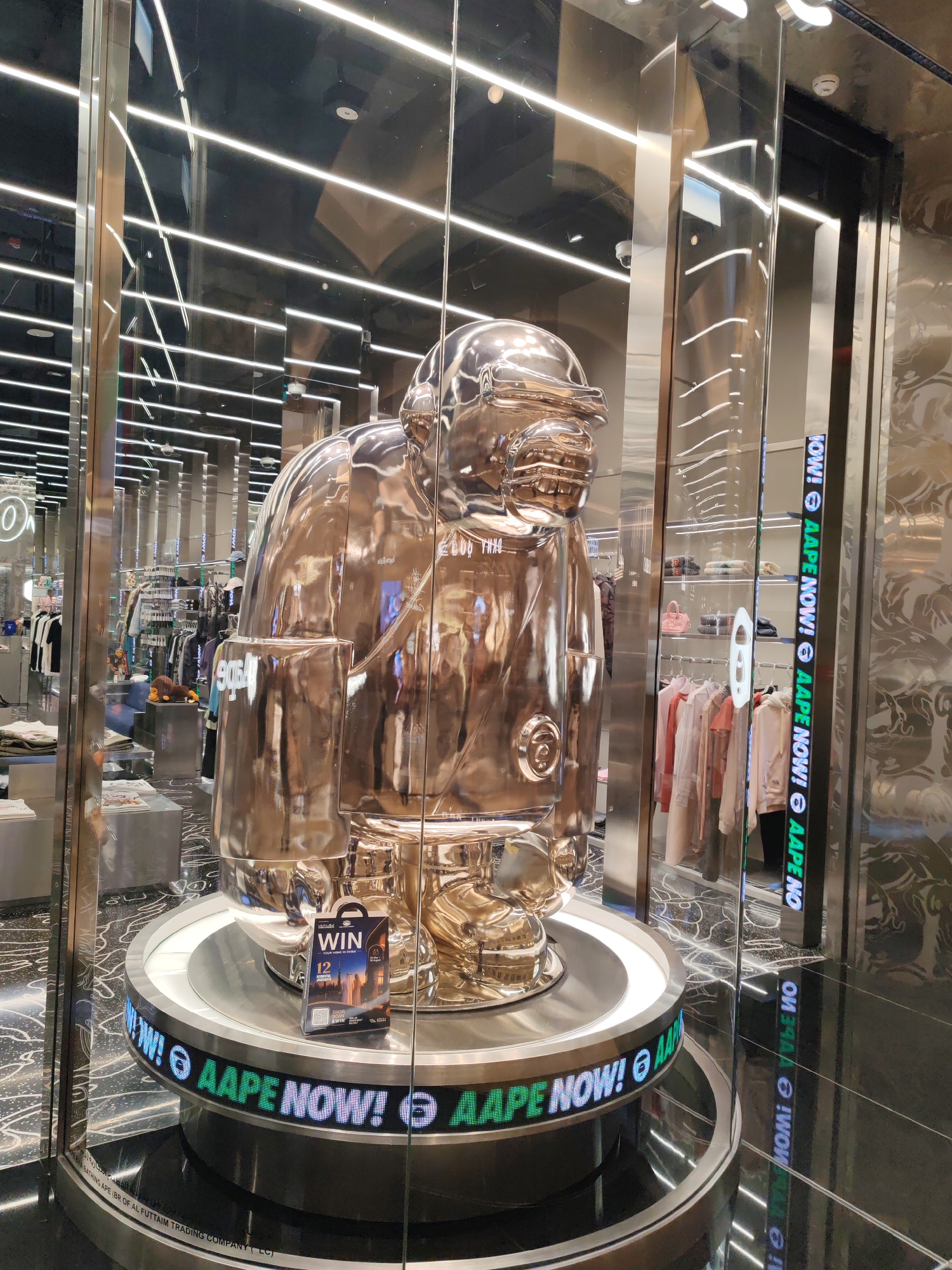 The image shows a large, shiny, silver-toned statue of a stylized ape, possibly a mascot, displayed in a glass-enclosed storefront. The statue is positioned on a circular platform with a digital display around its base that reads "AAPE NOW!". There is a promotional flyer on the platform to the right of the statue with the word "WIN" prominently displayed.
The background of the store is visible through the glass, showing racks of clothing, shelves with folded garments, and various decorative elements. The store appears to be a retail environment, likely a fashion boutique.
The lighting in the store is bright, with numerous linear fluorescent lights visible on the ceiling and reflected in the shiny surfaces of the statue and the store's interior. The flooring has a reflective, patterned design.
The scene is situated in a commercial area, and based on the text "AL FUTTAIM TRADING COMPANY (LLC)" on the base of the platform, the location is likely a retail space managed or owned by the Al-Futtaim Group. This group operates numerous retail brands and has a significant presence in Dubai, United Arab Emirates. Therefore, the image is very likely from a retail store in Dubai, UAE, possibly within one of the Al-Futtaim Group's retail destinations.