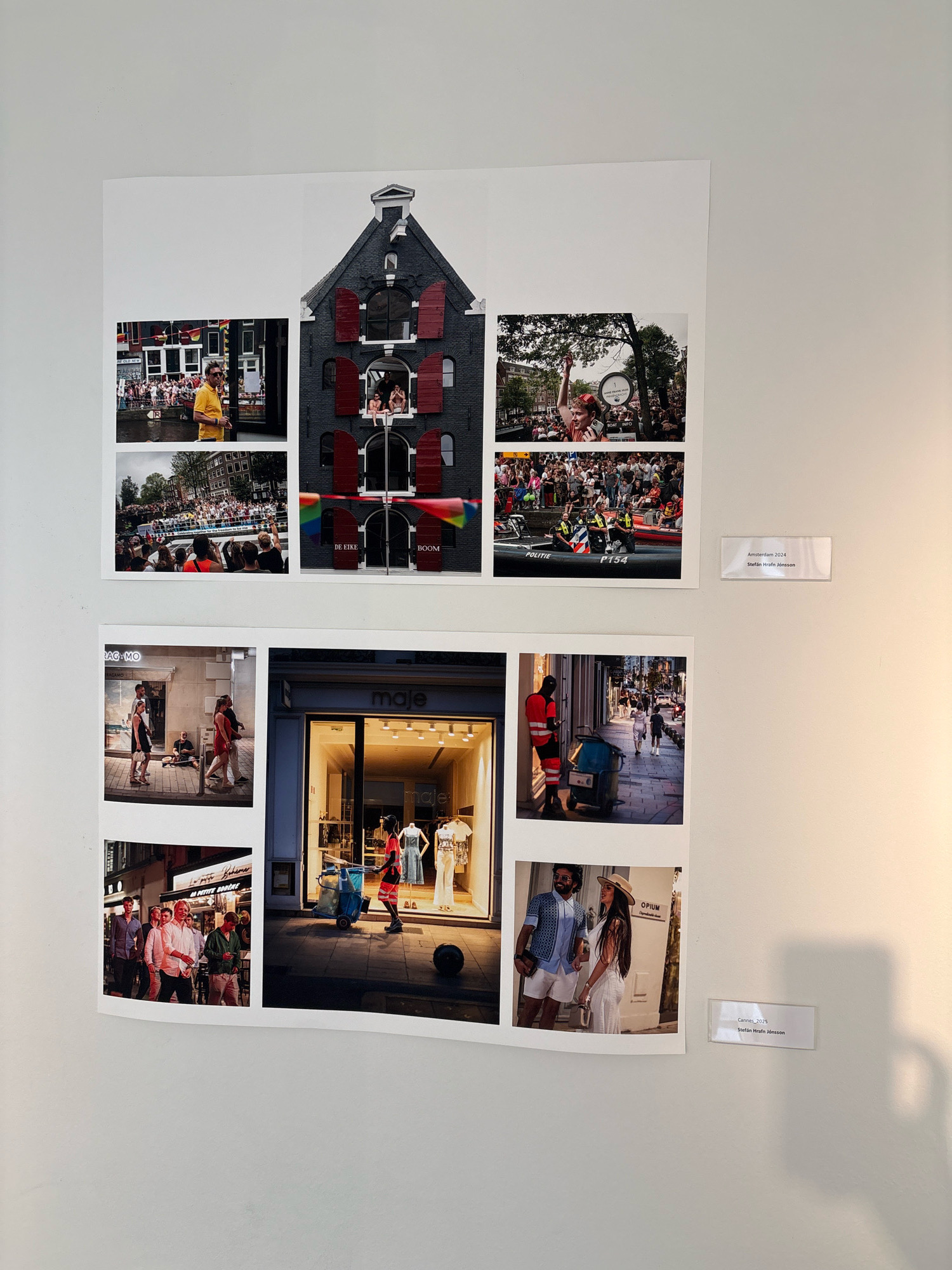 The image displays a wall with photographs mounted on it. The photographs are arranged in a grid-like structure.
In the upper left section, there are three photos. The top left photo shows a man in a yellow shirt standing next to a canal. Below him, a photo shows a canal boat filled with people. To the right of these two photos, a large photo depicts a dark building with red shutters and rainbow flags hanging from it. In the windows of the building, several people are visible.
In the lower section, there are four more photos. The bottom left photo shows a group of men walking on a street. To the right of that, a photo shows a storefront with mannequins inside. Below the storefront photo, on the left, is a photo of four people standing on a sidewalk. To the right of that, a photo shows a man and a woman posing for a photo on a street.
On the wall, to the right of the photographs, there are two small rectangular signs. The upper sign reads "Amsterdam 2024 Stefan Hrafn Jönsson". The lower sign reads "Cannes 2025 Stefan Hrafn Jönsson".
The background appears to be a light-colored wall, with some shadow cast on it from the artwork. The lighting suggests an indoor setting, possibly a gallery or exhibition space. There is no visual information in the image that suggests the location is Reykjavik, Iceland.