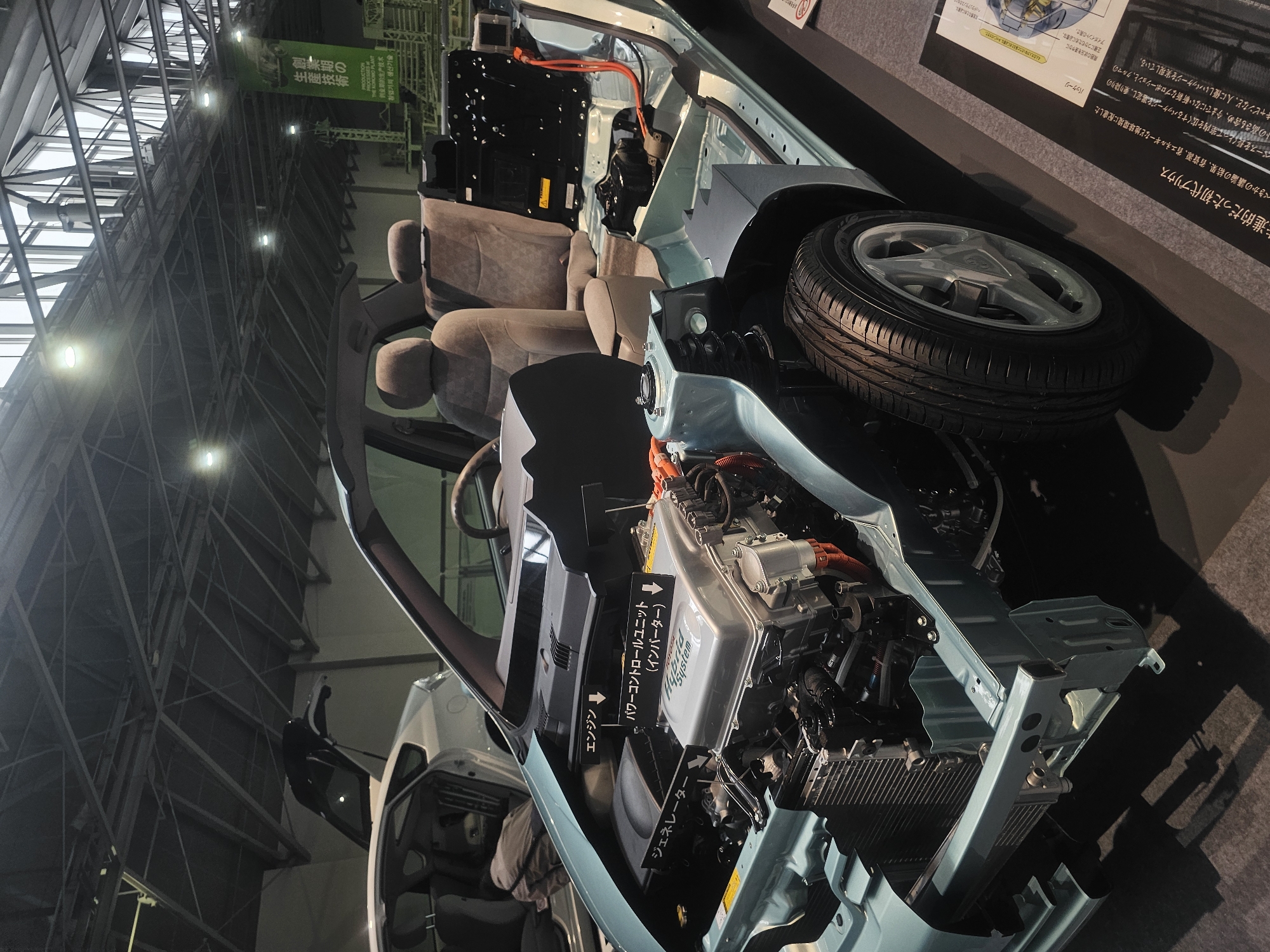 The image shows a cutaway display of a Toyota Prius hybrid vehicle, likely at an automotive exhibition or museum. The vehicle's interior and engine compartment are exposed, revealing the powertrain components, including the engine and generator, labeled in Japanese. The car is displayed indoors, with a high ceiling structure visible, suggesting a large exhibition hall. In the background, a green banner with Japanese text is hung, possibly indicating the exhibition's theme or sponsor. The overall scene depicts a static display of automotive technology. While the image is a detailed view of the vehicle, there is no specific indication of its location in Nagoya, Japan.
