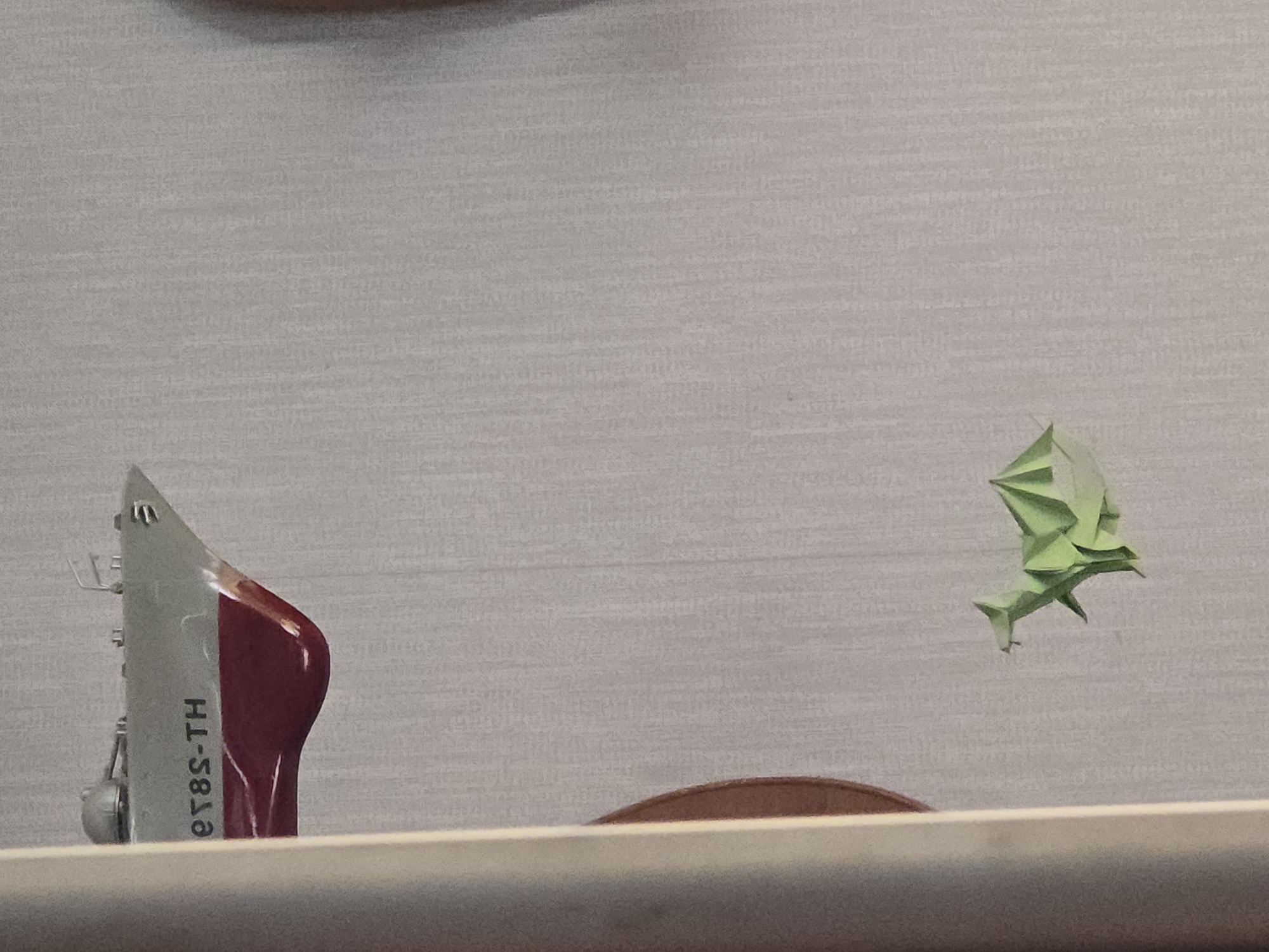 A model of a ship is visible in the upper left quadrant of the image. The ship is white and red, with "er8S-TH" written in black lettering on its side. In the lower center of the image, a green origami dragon is attached to a textured wall. The wall appears to be wallpapered. A portion of a cylindrical object, possibly a lamp or pillar, is visible on the left edge of the image, obscuring part of the scene.