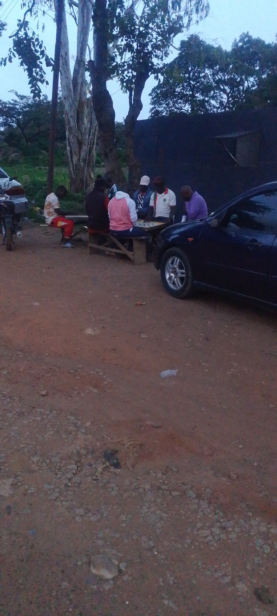 In an outdoor, dirt-ground setting, a group of six men are gathered around a table, some seated on benches, others on what appear to be makeshift seats. The scene is dimly lit, suggesting it is dusk or dawn. The men are engaged in an activity involving what appears to be a game board on the table. A dark colored car is parked to the right of the scene, and a motorcycle is visible to the left. Trees with sparse foliage and a utility pole are present in the background. The environment appears rural or on the outskirts of a settlement, with a dark, corrugated structure visible behind the trees.