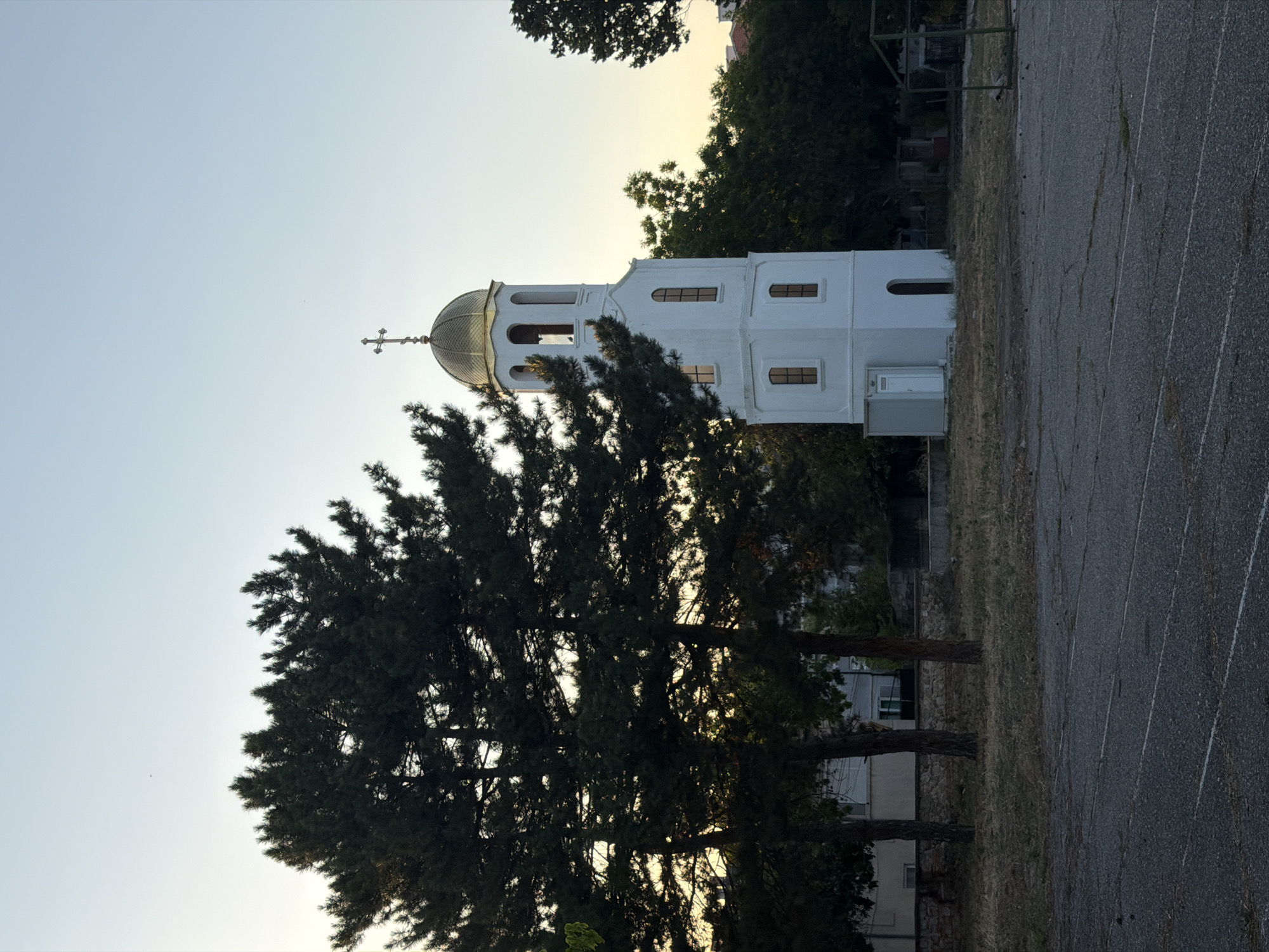 The image shows a white bell tower with a golden dome topped by a cross. The tower has several arched windows and a door at its base. It is located in the city of Svilengrad, Bulgaria. In the foreground, there is a paved area with white lines, resembling a sports track. Large pine trees are prominent on the left side of the image, with their branches partially obscuring the bell tower and a building behind them. The background consists of more trees and some residential buildings. The sky is clear and light blue, with a hint of yellow at the horizon, suggesting either sunrise or sunset. There are no people or animals visible in the image. The overall scene appears to be an outdoor, possibly public, space.
