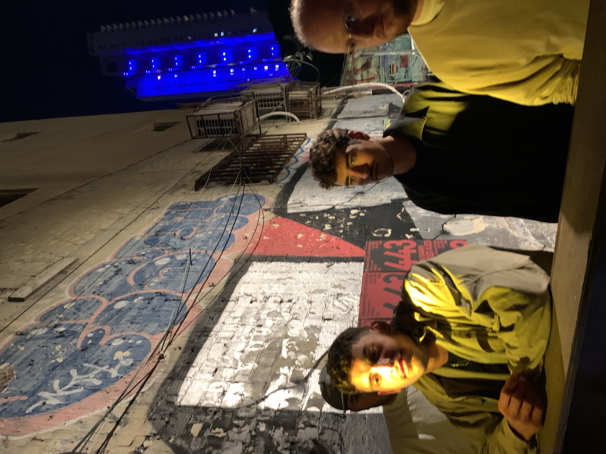 This image captures an urban night scene in Tel-Aviv, Israel, featuring three individuals against a backdrop of a graffiti-covered wall and a brightly lit building. The image appears to be taken from a slightly low angle or the subjects are looking down towards the camera, creating a unique perspective.
In the foreground, two men are clearly visible, both looking towards the camera. The man on the upper right wears a yellow long-sleeved top and glasses. Next to him, a younger man with dark, curly hair wears a black t-shirt; his face is highlighted by a warm, artificial light source. A third person, partially visible on the lower right, wears a light-colored (possibly yellow or olive green) jacket and also looks towards the camera, resting their hand on a dark ledge or railing.
The background reveals a tall building with striking blue lights illuminating its upper stories and details, standing out against the dark night sky. Below the building, a textured wall fills much of the frame. This wall is heavily adorned with various layers of street art and grime. Notable graffiti includes an abstract design in blue and pink hues, and another section with a red and black pattern featuring visible text fragments like "443" and "EFFECT". Several rusty fire escapes and multiple wires run vertically and horizontally across the wall.
The scene is lit by artificial lights, indicating nighttime. The faces of the individuals and sections of the wall are brightly illuminated, creating strong contrasts with the dark sky and shadowed areas.