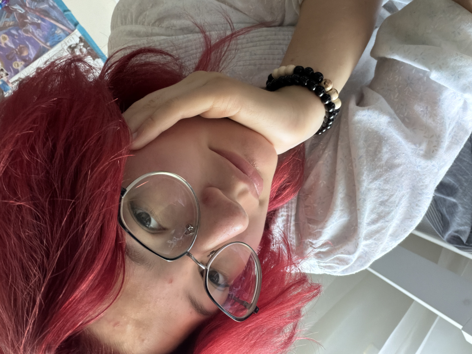 The image shows a close-up of a young person with vibrant red hair, wearing glasses and a white top. They are resting their head on their hand, and the background appears to be indoors with some shelves visible. The person is wearing two beaded bracelets on their wrist, one black and one with alternating black and cream beads. There is no visible indication of the city of Chelyabinsk, Russia, in the image.