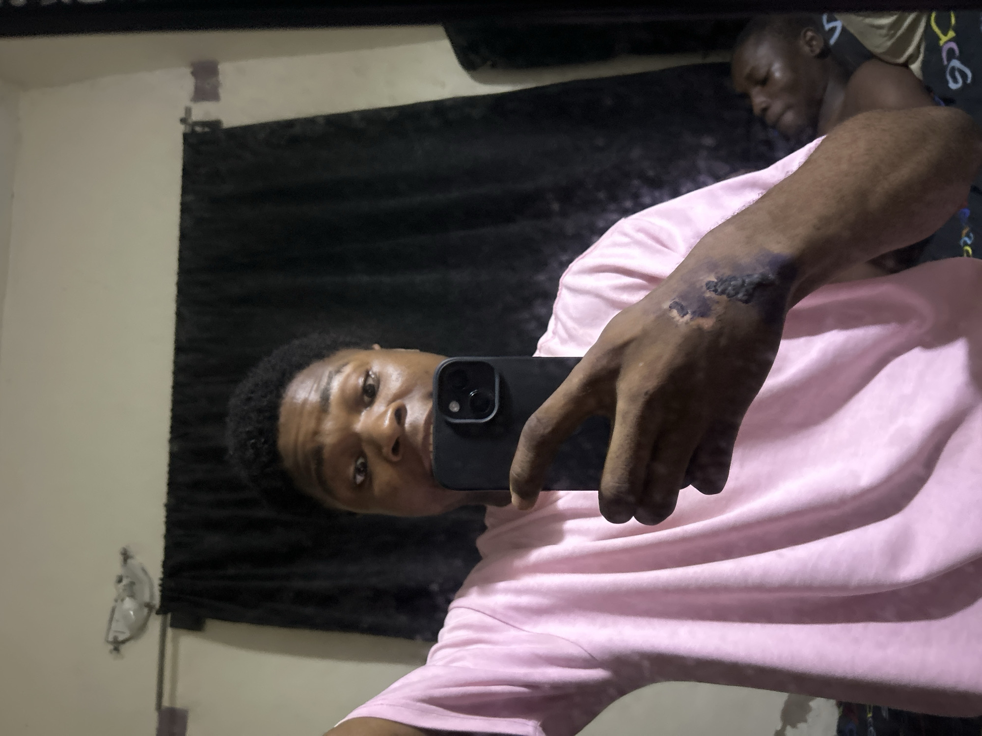 A young Black male, identified as the subject, is taking a selfie in a mirror. He is wearing a pink t-shirt and is holding a black smartphone in his right hand, which is raised to capture the reflection. His left arm is visible, and there is a dark, textured mark or injury on his forearm. In the background, reflected in the mirror, another individual, also Black, appears to be lying down, partially visible. The environment appears to be a room, with a dark curtain behind the subject and a light fixture on the wall. The overall lighting is dim. The scene is likely located in a residential setting in Ihiagwa, Nigeria.