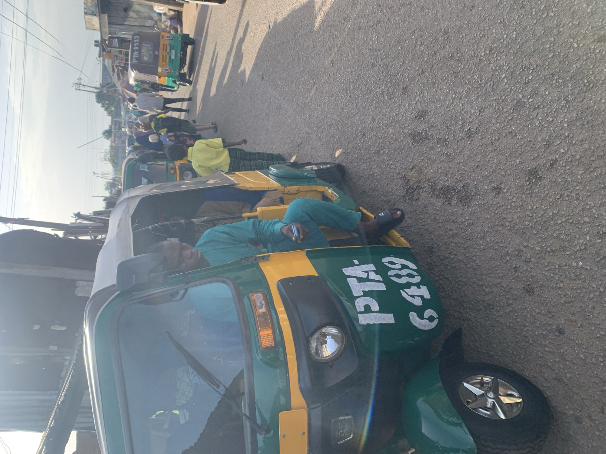 In Jos, Nigeria, a man in a teal outfit reclines in the driver's seat of a green and yellow auto-rickshaw. The auto-rickshaw, parked on a paved road, displays the text "PTA 6489" in white lettering. Behind him, a line of auto-rickshaws stretches down the street, suggesting a transport hub or a queue for passengers. Several people are visible in and around the other vehicles. The sun is high in the sky, casting long shadows from the vehicles and overhead wires, indicating it's likely midday. The scene appears to be a typical urban street in Nigeria, with buildings lining one side of the road and the general atmosphere suggesting a busy day.