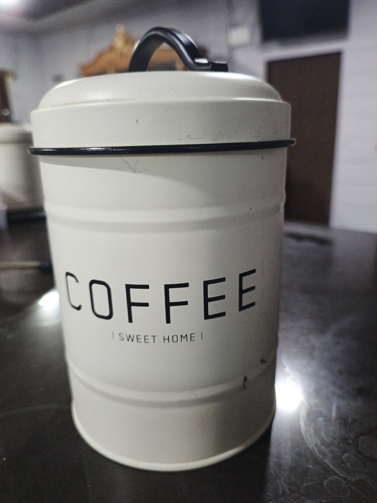 A white metal coffee canister with black lettering that reads "COFFEE" and "SWEET HOME" is positioned on a dark, reflective surface, possibly a table. The canister has a black handle on its lid. The background is blurred, showing hints of a room with a door and possibly furniture. The lighting suggests an indoor environment, and there are no people or animals present in the image. The specific location in Kelapapati, Indonesia, is not discernible from the visual information provided in the image.