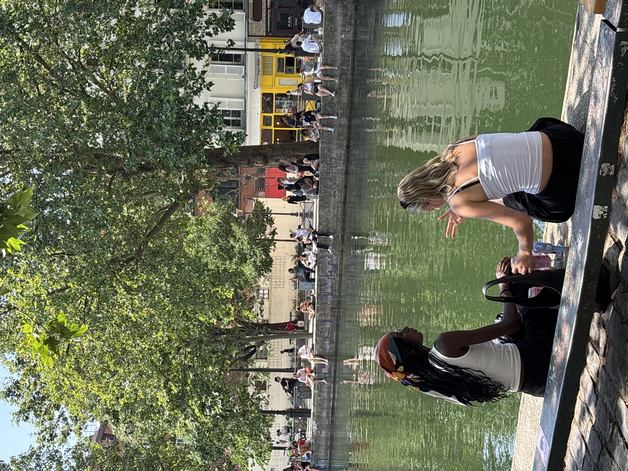 The image shows a sunny day along the Canal Saint-Martin in Paris, France. Two young women are sitting on the edge of the canal on a concrete ledge, facing the water. The woman on the left has dark skin, long dark braided hair, and is wearing a white tank top and a colorful headscarf. She is looking to her right. The woman on the right has blonde hair and is wearing a white tank top and black pants. She is facing the woman on the left and appears to be talking, with her hands raised. A black handbag is resting next to the woman on the left. On the other side of the canal, a line of people are sitting and standing along the edge, some under the shade of large trees. Buildings with yellow and white facades line the canal in the background. The water of the canal is a vibrant green and reflects the sunlight and surrounding trees and buildings.