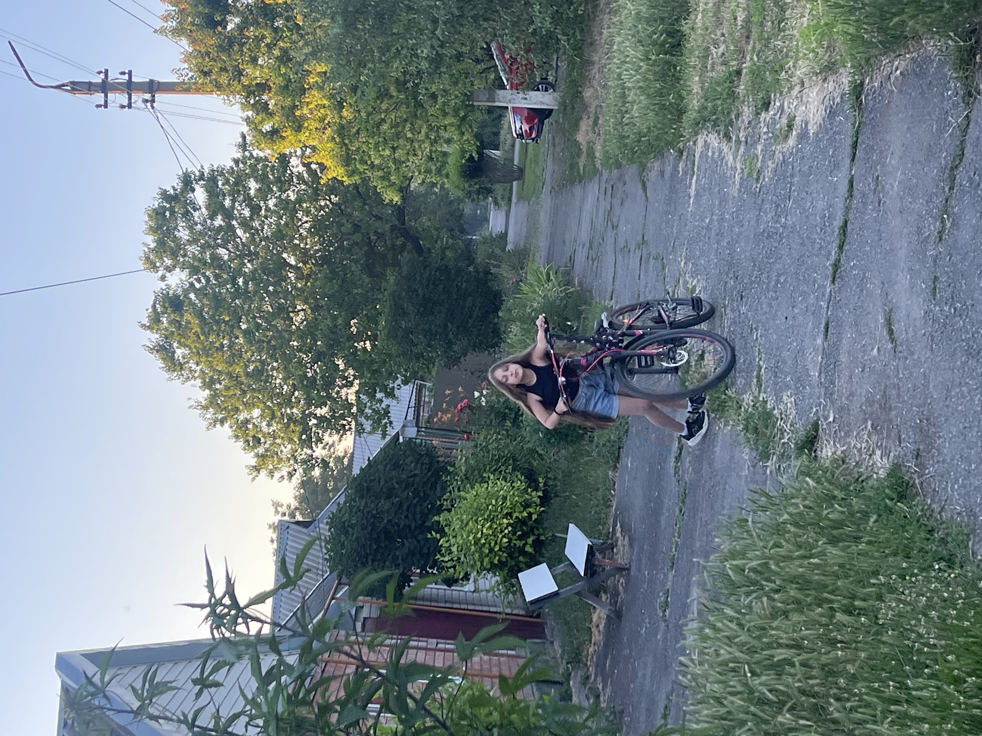 A young woman is standing on a paved pathway, holding onto a bicycle. She is wearing a black tank top and denim shorts. The bicycle has a pink frame and black tires. The pathway is lined with lush green grass and tall trees with green foliage. In the background, there are several houses and a utility pole with wires running across the frame. The sky is a clear blue, suggesting it is daytime. The scene appears to be in a residential area. The woman's expression is relaxed.