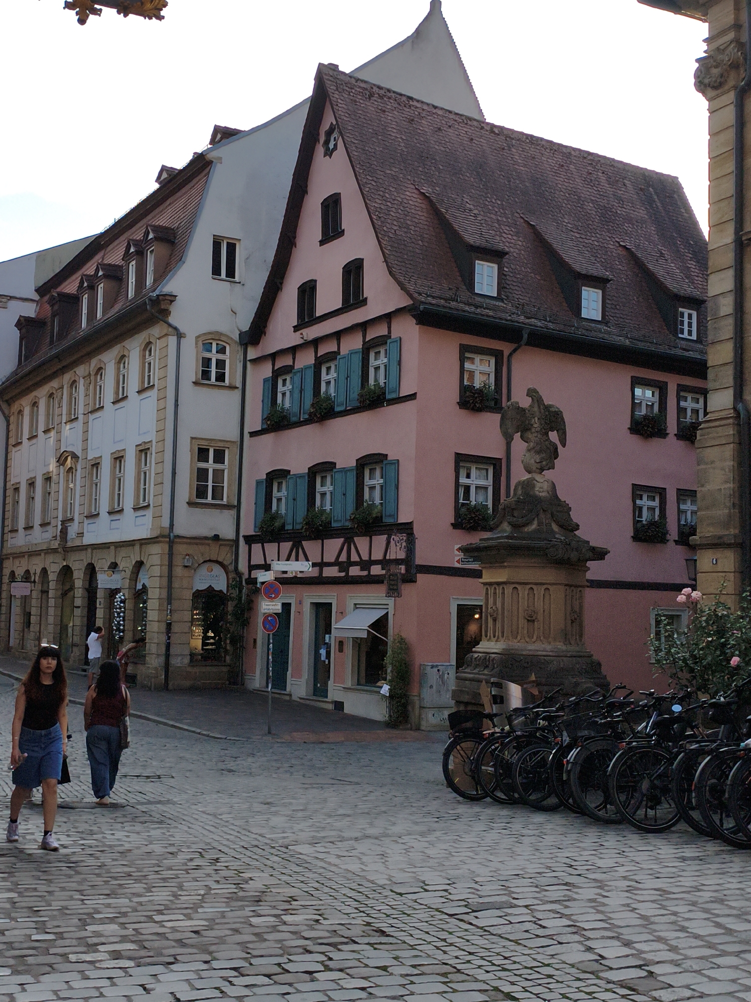The image depicts a street scene in Bamberg, Germany, specifically in the vicinity of the Maxplatz. In the foreground, two women are walking on a cobblestone street. The woman on the left is wearing a black tank top and a denim skirt, while the woman on the right is wearing a maroon t-shirt and blue jeans. Behind them, a row of bicycles is parked along the sidewalk. The street is lined with historic buildings, including a light blue building with arched ground-floor windows and a pink building with dark trim and teal shutters. A stone fountain with a bird sculpture stands in front of the pink building. The architecture suggests a European city with well-preserved historical structures. The lighting indicates it is late afternoon or early evening, with soft shadows cast across the street.