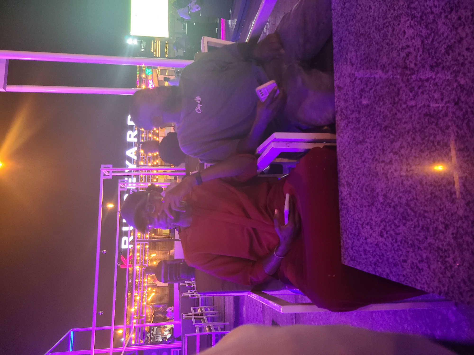 Here is a description of the image:
The image shows two people seated at an outdoor venue. The person in the foreground is wearing a red outfit. The person behind them is wearing a gray t-shirt and holding a smartphone. The background is illuminated with bright purple and orange lights. A sign that reads "RIV YARD" is visible in the background. Based on the information available, the venue is located in the city of Gura Top, Nigeria. There are tables and chairs behind the two people. The lighting suggests it is nighttime.