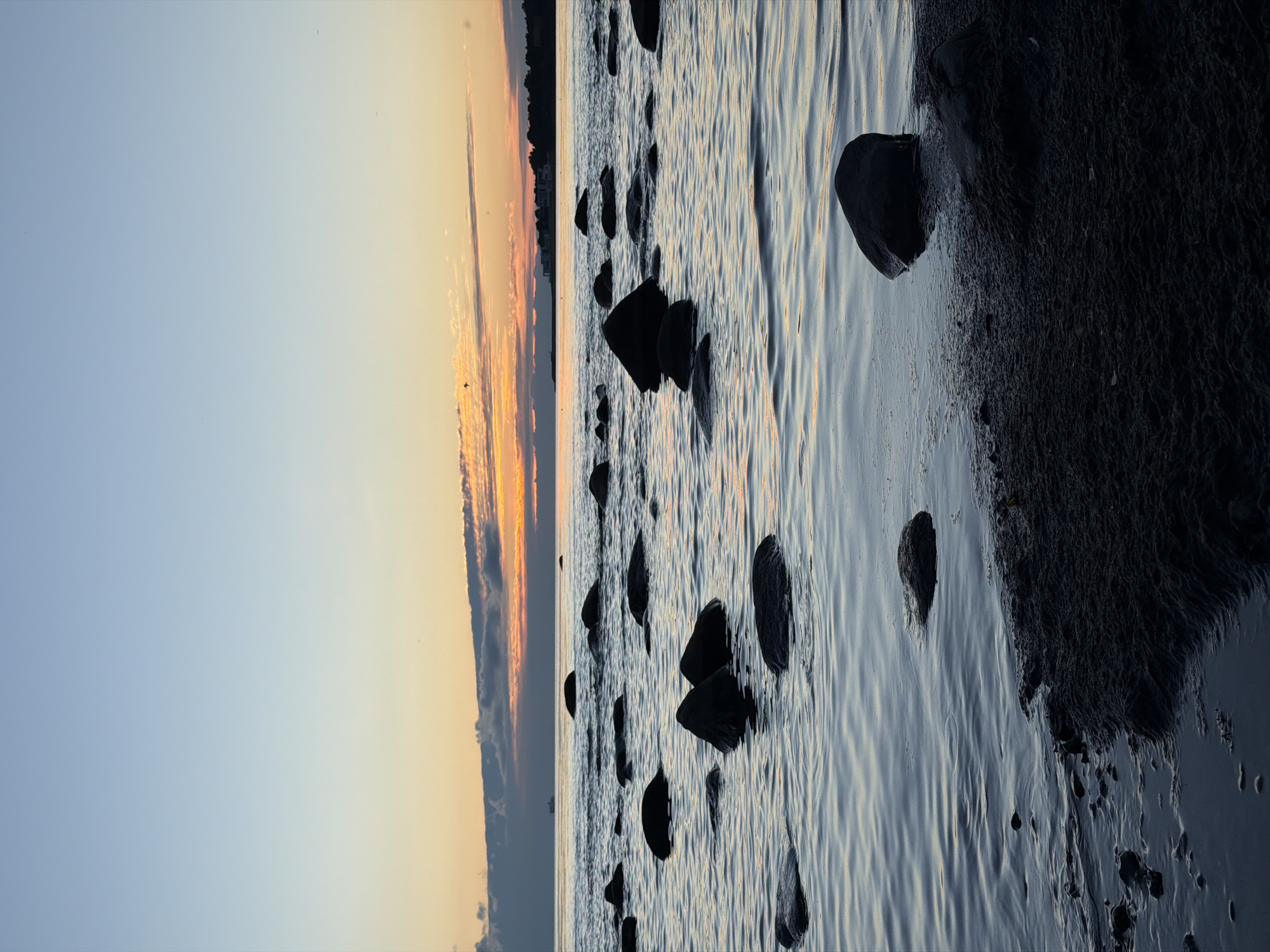 This image depicts a serene sunset over the Baltic Sea in Haabneeme alevik, Estonia. The sky transitions from a pale blue at the top to soft oranges and pinks near the horizon, where scattered clouds are illuminated by the setting sun. A distant ship is visible on the water. The foreground is dominated by a calm sea with numerous dark, rounded rocks emerging from the water and along the sandy shoreline. The water exhibits gentle ripples, reflecting the muted colors of the sky. The lighting suggests the scene is captured during the twilight hours. There are no people or visible actions taking place.