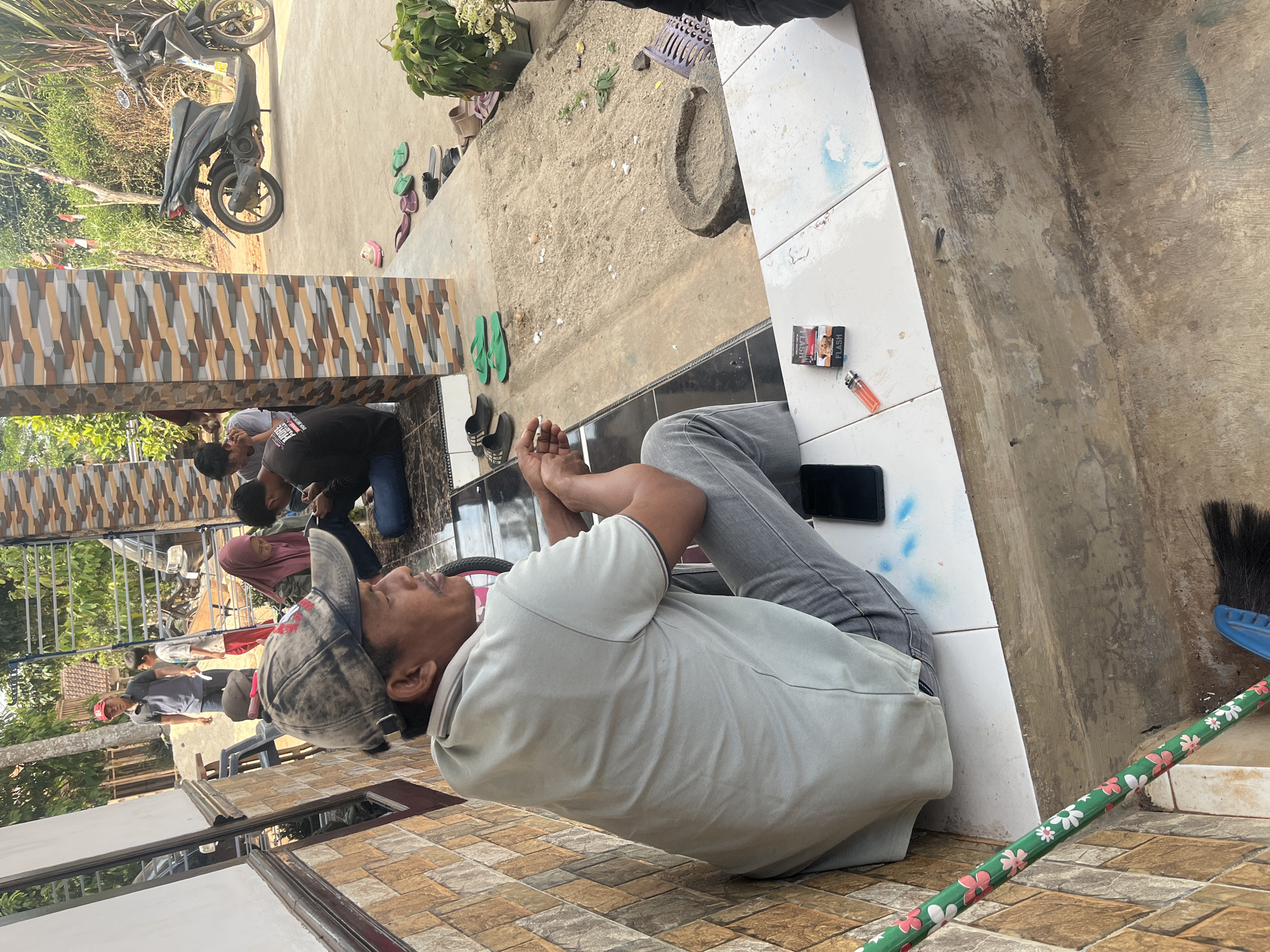 This is a medium shot photograph taken in Karang Sari, Indonesia. In the foreground, a man wearing a light grey polo shirt, grey jeans, and a dark baseball cap sits on a tiled step. He is looking to the right and holding a cigarette. Next to him, on the step, are a pack of cigarettes and a lighter. A smartphone is also visible on the step. To the left of the man, a broom with a green handle decorated with pink and white flowers is leaning against a tiled wall.
In the background, several other people are seated or standing. A woman in a pink hijab and a dark shirt is seated. Another person in a black shirt is seated and looking at a phone. Two children are standing near a man wearing a red cap and a dark shirt. A black scooter is parked on a concrete surface behind the people. The environment appears to be a residential area with some vegetation and a tiled building.