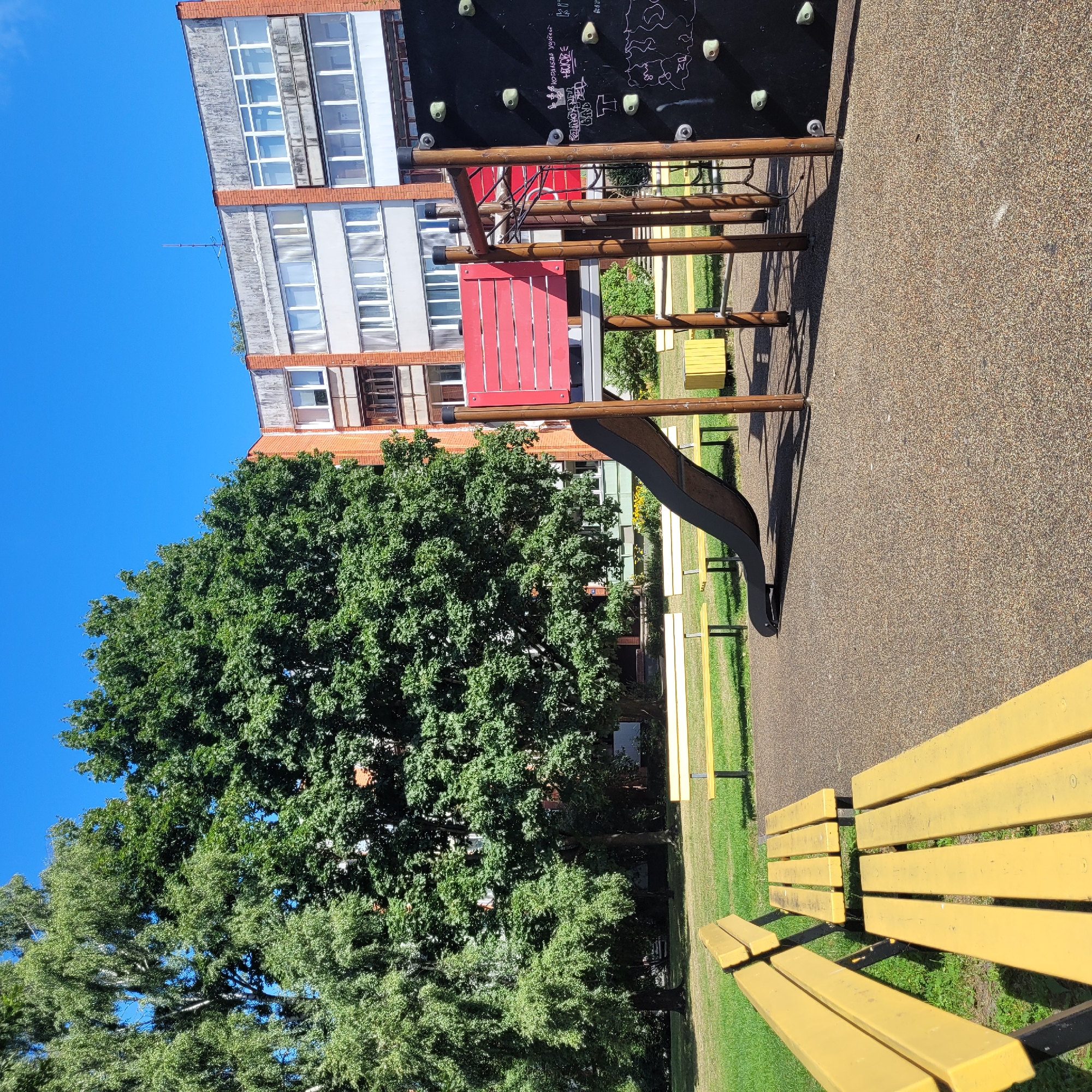 This image depicts a playground located in the city of Jūrmala, Latvia. The playground features a slide, a climbing wall, and several yellow park benches. The ground surface of the playground is composed of a brown, pebbled material. Behind the playground, a large tree with lush green foliage dominates the scene, with a multi-story residential building visible through the trees. The sky is a clear, bright blue. The overall environment suggests a sunny day.