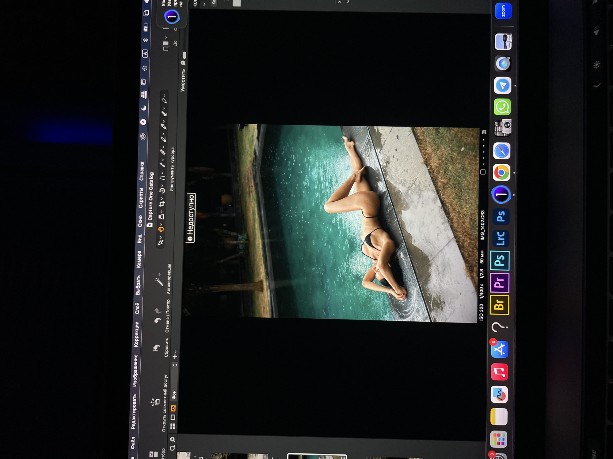 The image shows a woman in a black bikini lying on the edge of a pool. The pool is surrounded by greenery and a concrete edge. The image is displayed on a MacBook Pro screen, within the Capture One photo editing software. The computer screen also shows the dock and the menu bar with various application icons and settings.