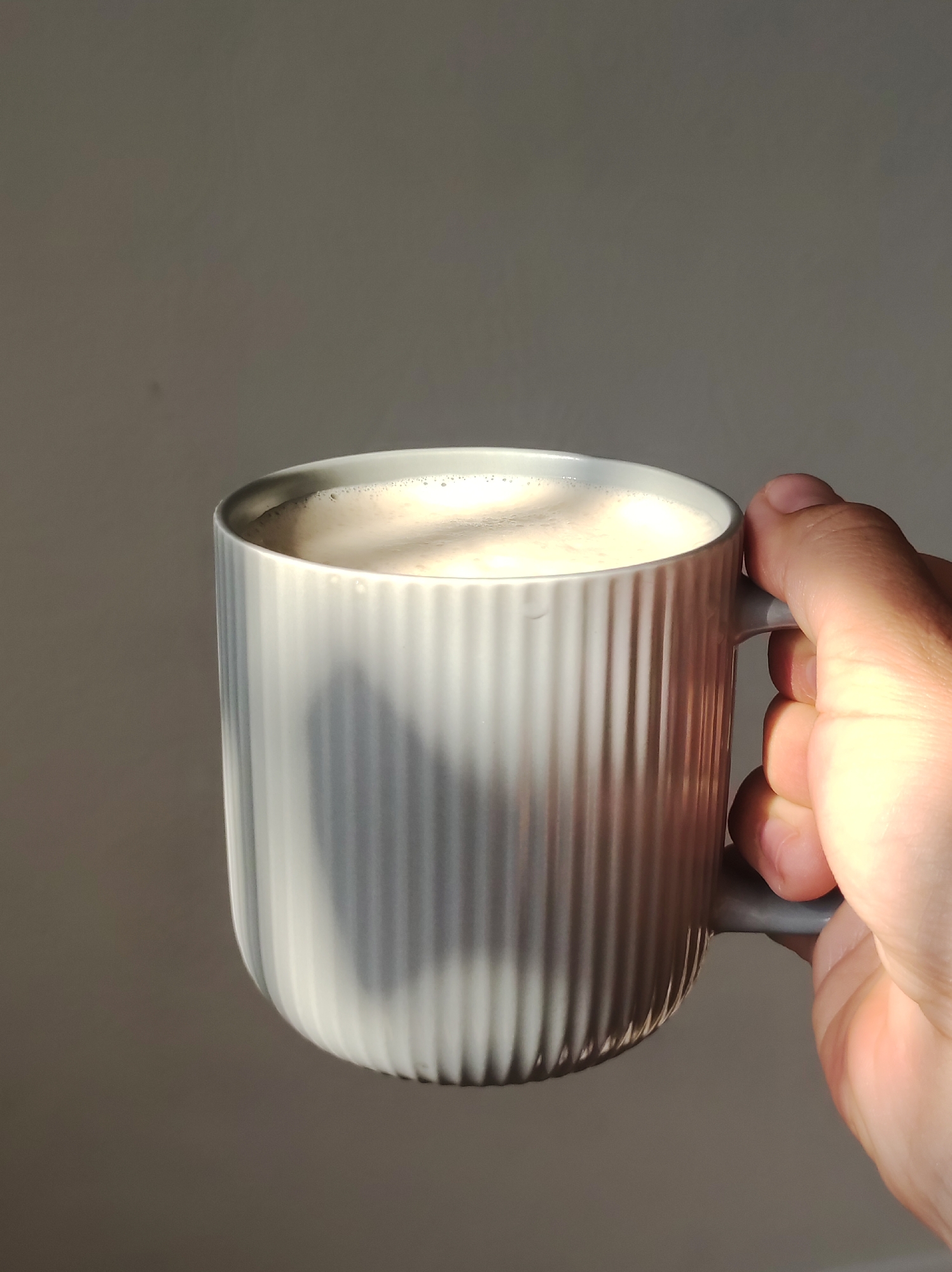A hand is holding a ribbed, light gray mug filled with a foamy beverage, possibly coffee or tea. The mug is positioned in the right side of the frame, and the hand is gripping its handle. The background is a plain, gray wall. Sunlight casts a shadow on the mug and illuminates the hand. The scene appears to be indoors.