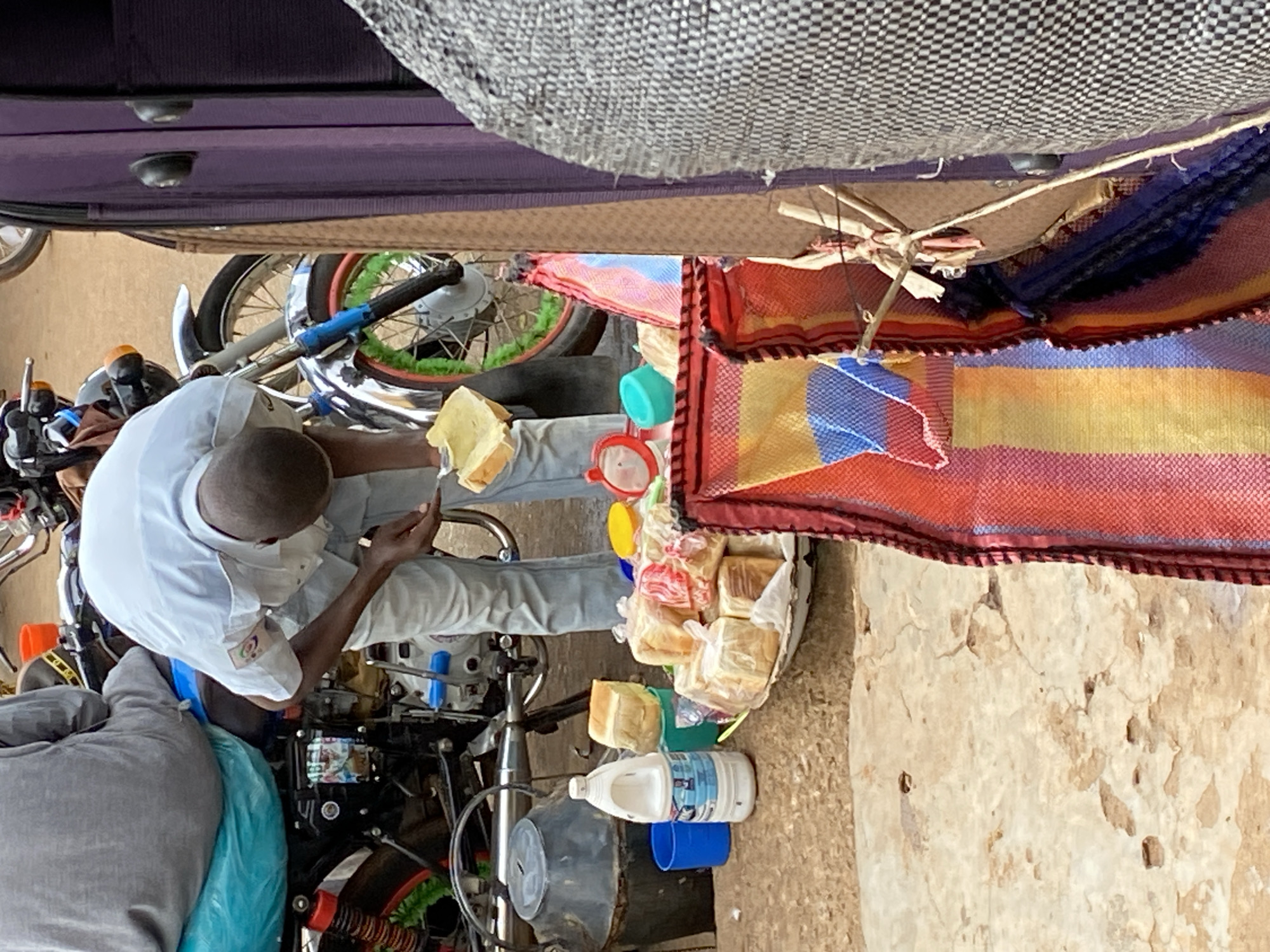 An individual wearing a white shirt and light-colored jeans is seen on a motorcycle in the city of Bukuru, Nigeria. The motorcycle is laden with loaves of bread, some of which are individually packaged in clear plastic bags. There's also a white plastic jug (possibly containing water or milk) and a blue plastic cup. The motorcycle is parked on a light-colored surface, possibly a concrete or dirt area. Above the motorcycle, there's a colorful woven fabric hanging, as well as what appears to be the undercarriage of a vehicle.