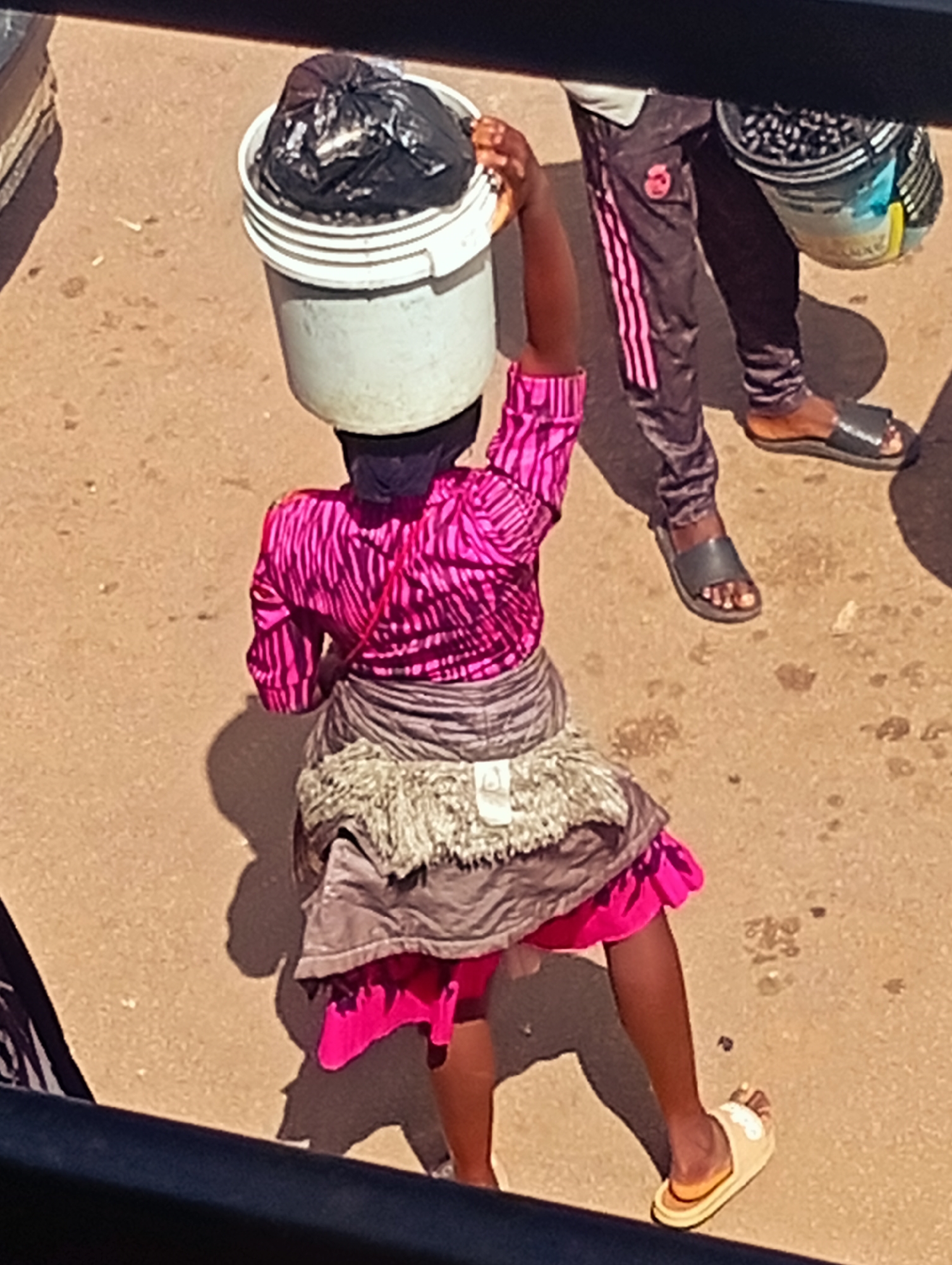 In Bukuru, Nigeria, a person carries a bucket of black plastic bags on their head. The person is wearing a vibrant pink and black striped shirt, a gray fringed wrap, and a bright pink skirt. They are also wearing sandals. To their right, another person is visible, wearing dark pants with pink stripes and black sandals. The scene appears to be outdoors on a paved surface.