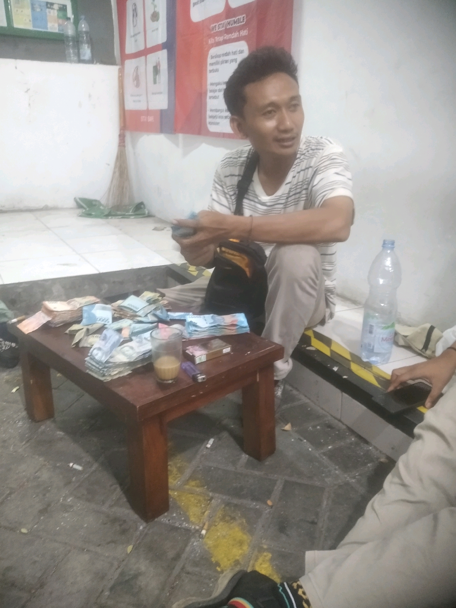 The image depicts an indoor scene, possibly a community hall or a casual meeting space, in Cilegon, Indonesia. A man in a striped t-shirt and light-colored pants is seated, holding a stack of Indonesian Rupiah banknotes in his hands, appearing to count or sort them. In front of him, on a low wooden table, is a substantial amount of cash, a glass with a light-colored liquid, a pack of cigarettes, a lighter, and some scattered cigarette butts on the floor. A partially filled bottle of water is visible to the right of the man, and another person's legs are visible in the lower right corner, holding a smartphone. The walls are white, with some posters or notices displayed. The floor is tiled and appears somewhat dirty with discarded cigarette ash and debris. The overall atmosphere suggests a casual gathering or a transaction taking place.