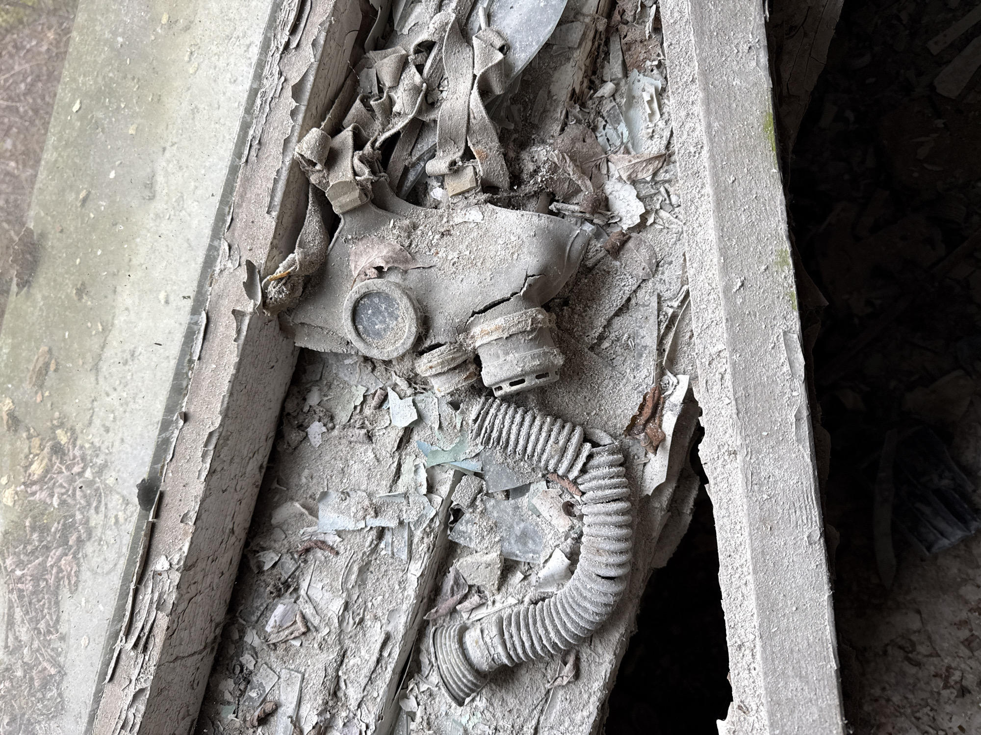 The image depicts a deteriorated gas mask resting amidst rubble and debris, possibly within a dilapidated structure. The mask's rubber components show signs of age and decay, covered in dust and debris. It is positioned within the confines of a window frame or a similar architectural element. The environment indicates an abandoned state, with broken pieces of glass and fallen plaster and other materials scattered around the gas mask. The context suggests a setting reminiscent of an abandoned building, the abandoned city of Prypiat in Ukraine being a likely location.