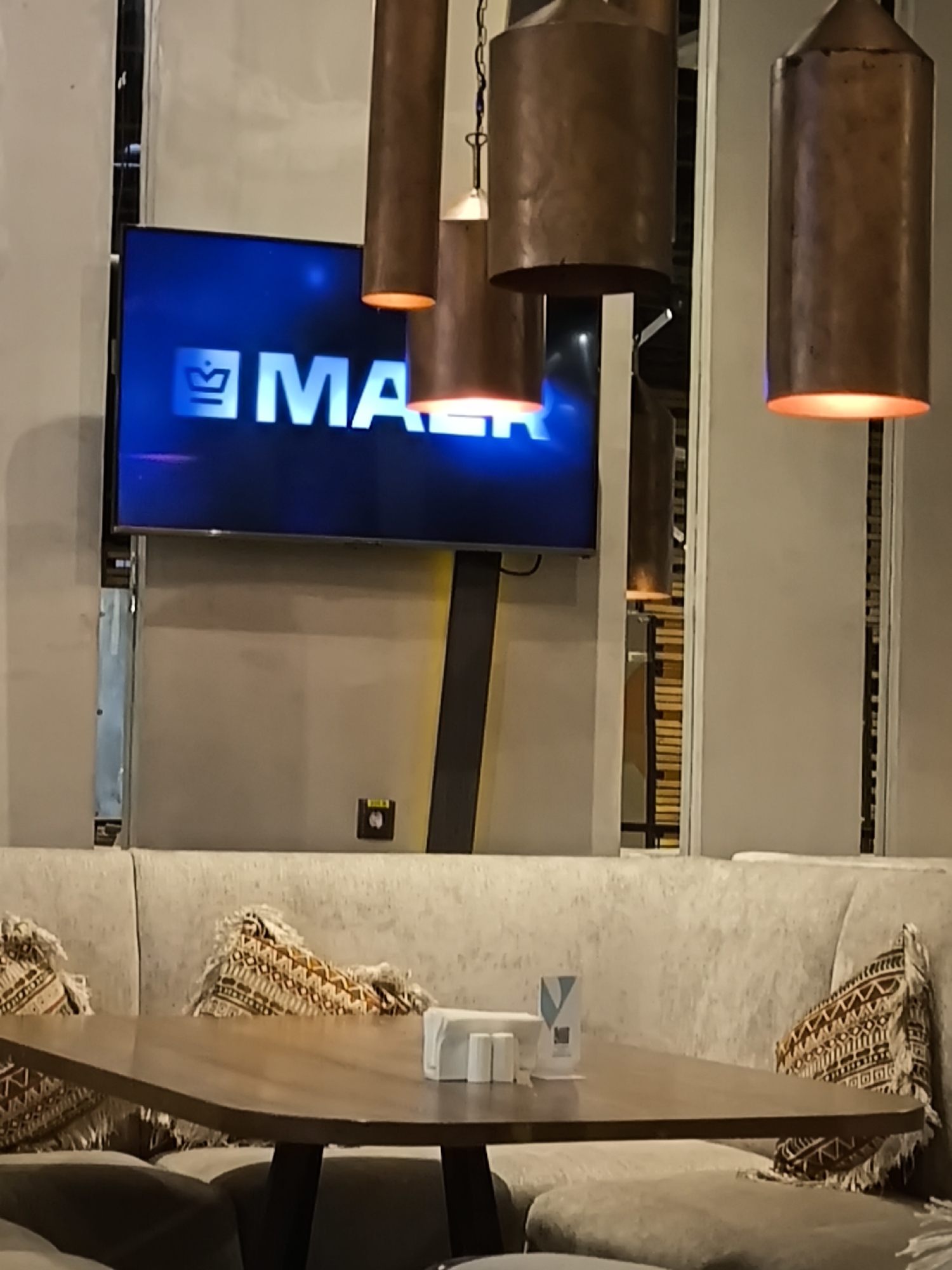The image depicts a dining area, likely in a restaurant, with a focus on the table and seating arrangement.
The foreground features a dark-legged wooden table, holding a napkin dispenser and salt/pepper shakers. Around the table is a built-in sofa-style seating area upholstered in a light beige fabric, adorned with patterned pillows.
Above the table, several copper-colored cylindrical pendant lamps hang at varying heights, casting a warm light downwards.
In the background, a large flat-screen television displays the word "MASK" in bold, white letters on a blue screen. The wall behind the seating is a light beige, and there appears to be a window or opening to another room filled with vertical wooden slats.
The image offers no explicit indication of Khimki, Russia.