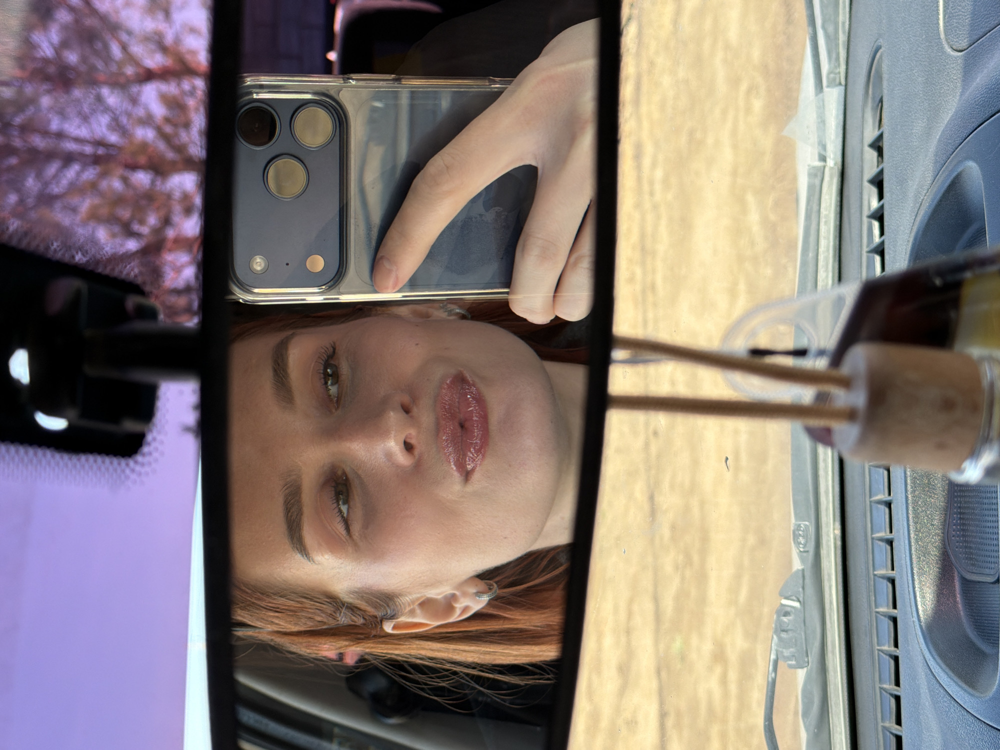 The image shows a reflection in a car's rearview mirror. A person with reddish-brown hair is visible in the mirror. They are holding a phone with a clear case, obscuring part of their face. The phone has multiple camera lenses. The mirror reflects the interior of the car, including the dashboard and a small air freshener with wooden sticks. Outside the car, blurry trees and foliage are visible. The image was most likely taken while the car was parked or stationary, but there is no indication of location, including Chaikovsky, Russia. There is no additional information available to confirm any location.