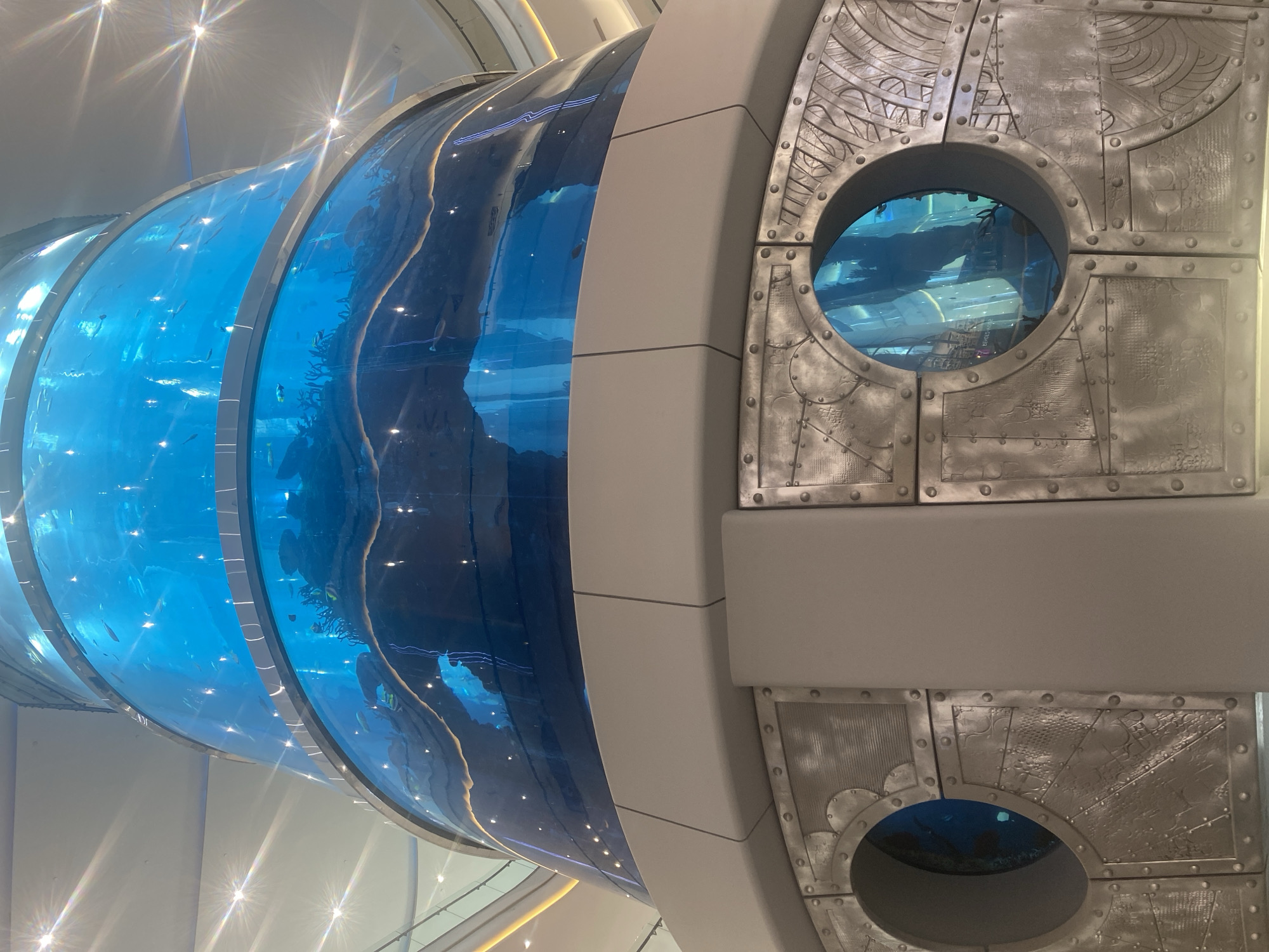 The image depicts a large, cylindrical aquarium filled with blue water and several fish, located in Moscow, Russia. The aquarium is segmented, with a wider, metallic base featuring circular windows. The upper section of the aquarium is transparent, allowing a view into the aquatic environment. The ceiling above is illuminated with star-shaped lights. The overall scene suggests an indoor public space, possibly a shopping mall or entertainment venue.