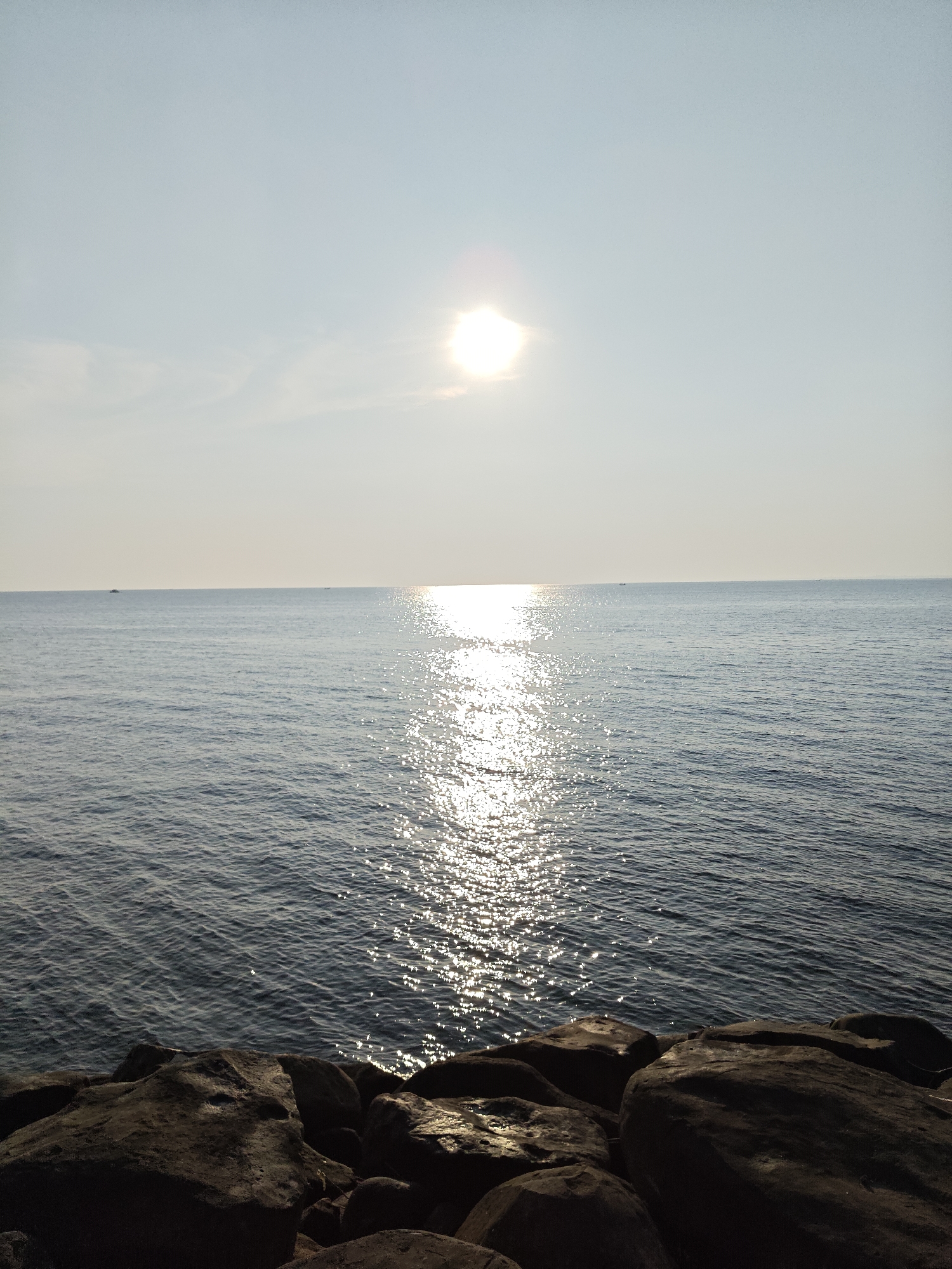 The image depicts a seascape at dusk or dawn, with the sun positioned high in the sky, casting a glare across the water's surface. The foreground features a pile of dark, large rocks. The water stretches to the horizon, and tiny, dark, undefined objects that may be boats are visible in the distance. The sky is a pale blue, slightly hazy. The location is likely a coastal area in Lhokseumawe, Indonesia, considering the latitude. No people are visible in the frame.