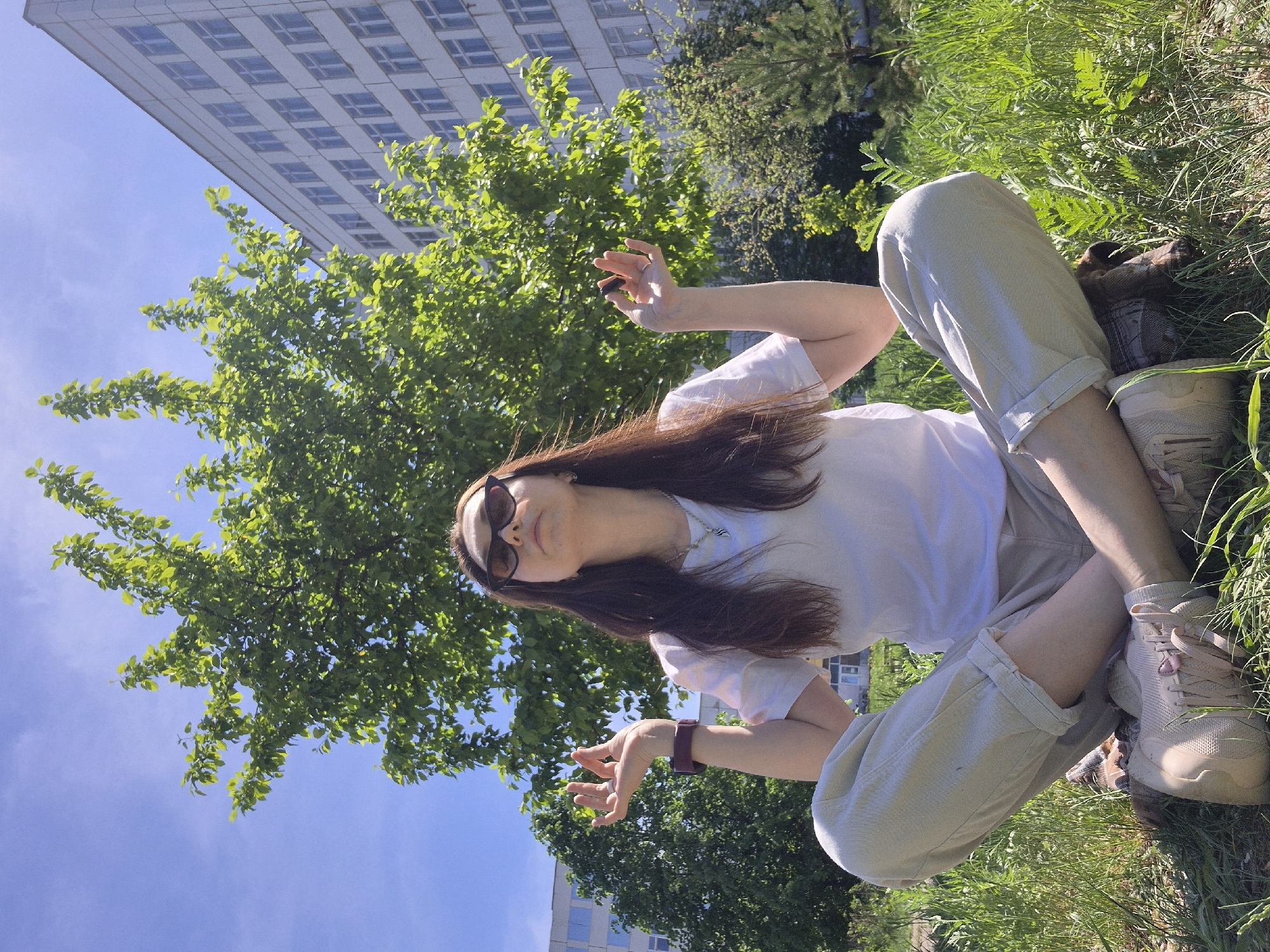A woman is sitting cross-legged on grass. She is wearing a white t-shirt, light-colored pants, and sunglasses. Her hair is long and brown. A tall building and trees are visible in the background. The location appears to be urban, possibly a park or green space within a city. The woman's arms are raised, with her hands open. She is holding a dark-colored rectangular device in one hand. She is also wearing a dark wristwatch.