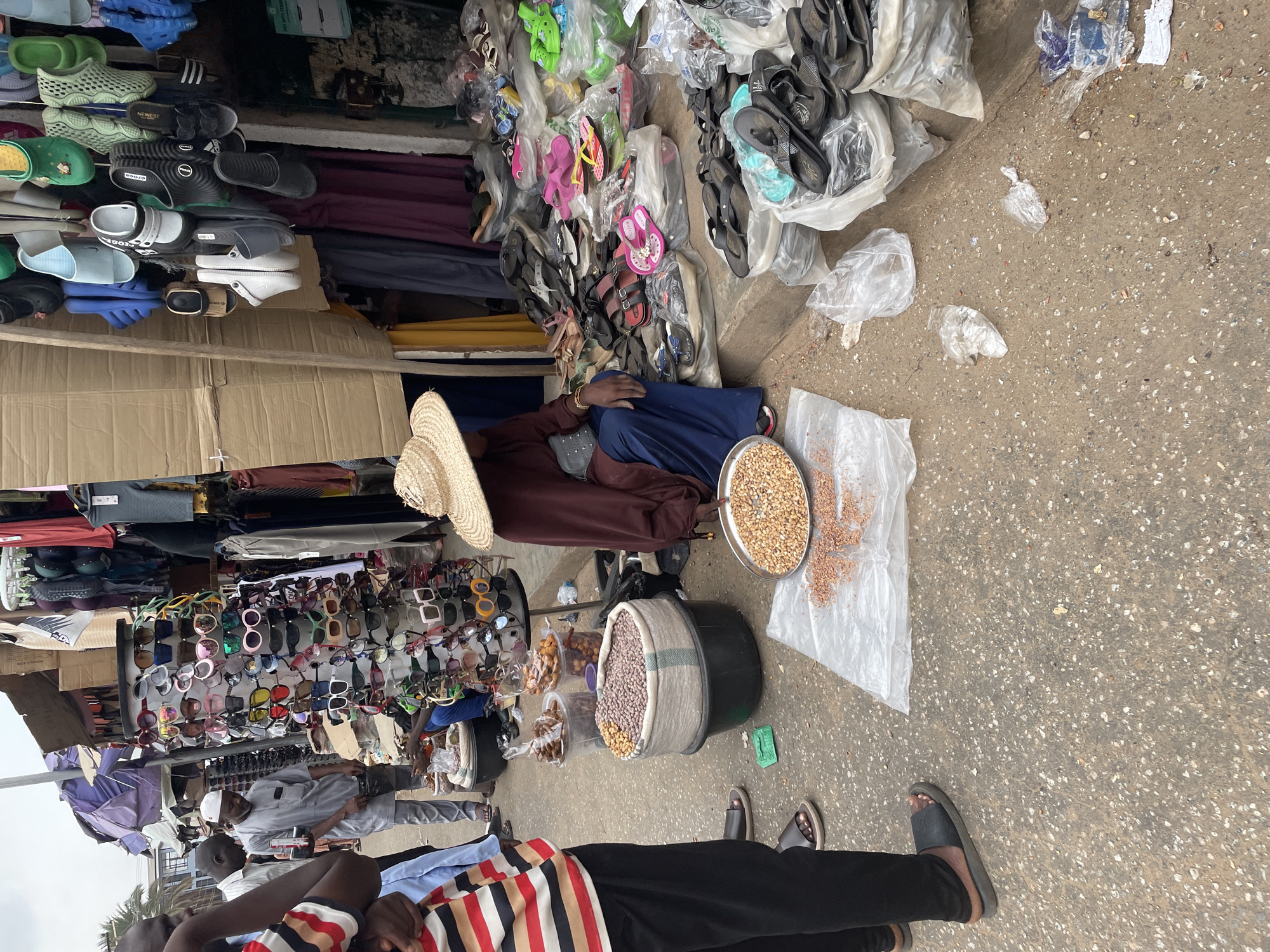 This media file captures a vibrant, bustling outdoor street market scene in Maiburiji, Nigeria, during the daytime. **Subjects:** Several people are visible in various roles. In the foreground, a person is seen from the chest down, wearing a striped shirt (red, black, white) and black trousers, with dark sandals on their feet. Another central figure is a vendor, sitting on the ground wearing a large straw hat, a brown long-sleeved top, and blue trousers. This vendor is surrounded by food items for sale. Further back, other individuals, likely shoppers or additional vendors, are standing and milling about. One man in a light grey outfit and a white cap is visible near a display of sunglasses. **Objects:** The market is densely packed with merchandise. On the left side, numerous pairs of footwear are displayed, including sandals, slides, and Crocs-like shoes, some stacked on shelves, others piled in clear plastic bags. Visible brands or labels include "CROCS" and "NEWEST," with a partial "adidas" logo on one shoe. To the right, more sandals and colorful flip-flops are also piled or bagged. A large rack densely packed with various styles of sunglasses is prominently displayed. The sitting vendor has a large round metal tray filled with light-colored grains or nuts, with some spilled onto a white plastic sheet on the ground. A large black bucket, partially lined with fabric, also contains a mix of grains or nuts, and smaller clear plastic containers hold other snack items. Cardboard boxes are used as part of the market stall structure. The ground is a speckled concrete or asphalt surface, scattered with plastic bags and small pieces of litter. **Scene/Setting:** The image depicts an informal outdoor market stall, likely situated along a street or pathway. The setting is somewhat cluttered, indicative of a busy commercial area. The overall atmosphere appears active and functional, typical of local commerce in Maiburiji. **Activity/Event:** The scene depicts a typical day in a bustling street market, where vendors are selling a variety of goods, including footwear, accessories like sunglasses, and food items such as nuts and snacks. People are engaged in buying, selling, or browsing. **Notable details:** The bright and even lighting suggests it is daytime, possibly late morning or early afternoon, with clear weather. There are no strong shadows, indicating either an overcast sky or midday sun. The market setup is rudimentary, relying on simple shelving, ground displays, and plastic bags for