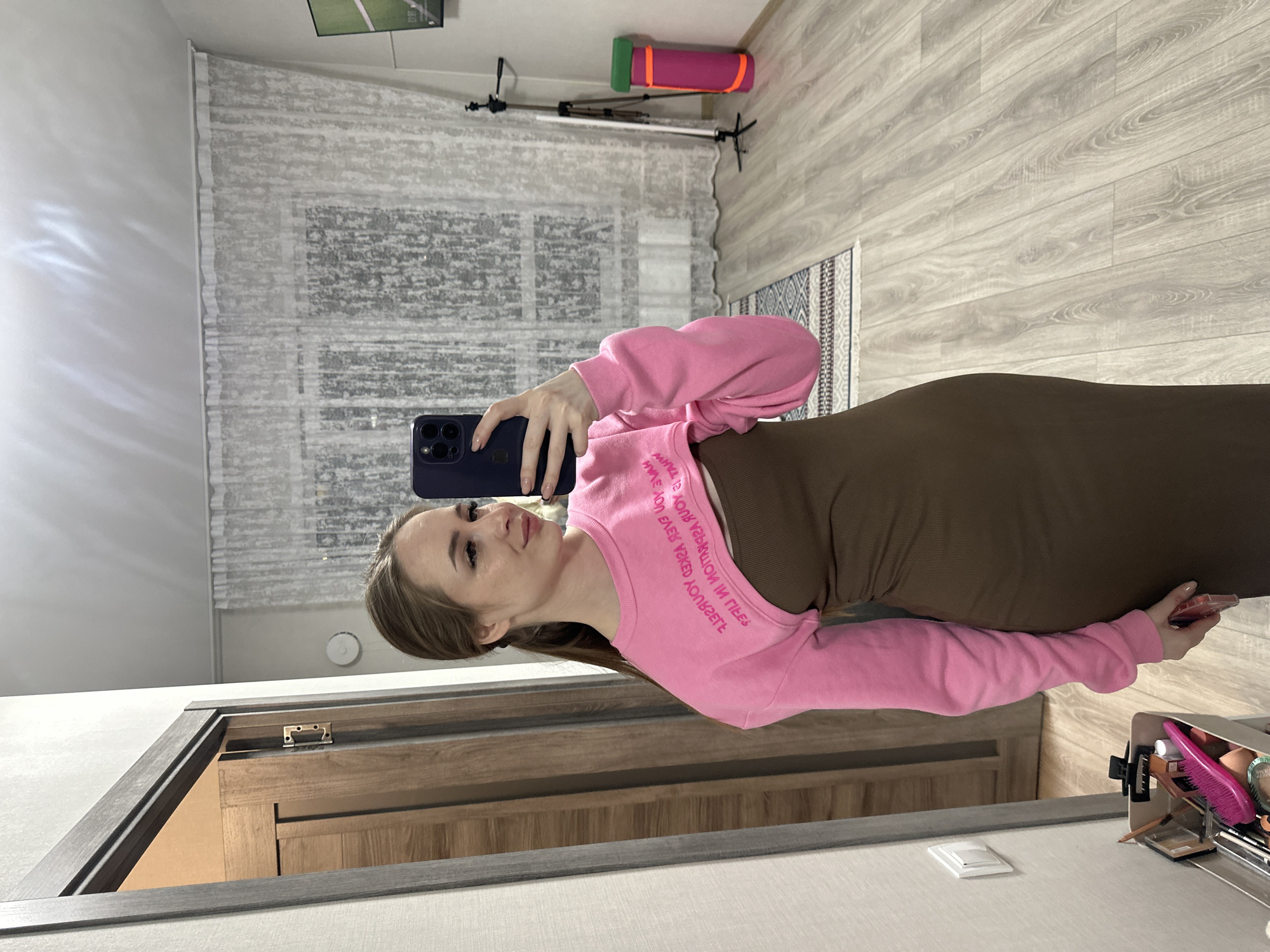 A young woman is captured in a mirror selfie, standing upright in an indoor setting. She has light brown hair and is wearing a vibrant pink long-sleeved cropped top over a dark brown, form-fitting midi-length dress. She holds a dark-colored smartphone in her left hand, using it to take the photo in the mirror. Her expression is serene, with a subtle smile.
The background reveals a room with light-colored wooden laminate flooring, accented by a small patterned rug in blue, white, and brown tones. The walls are a light, neutral color. Behind the woman, sheer, textured curtains cover a window, letting in diffused light.
Various objects are visible in the reflection and directly in the room. A rolled-up green and pink yoga mat rests on a shelf or ledge, next to a black tripod stand. On the right side of the mirror, a vanity or shelf displays cosmetics, including a bright pink hairbrush, reflecting prominently. A wooden door frame and part of a wooden door or cabinet are also visible in the mirror's lower reflection. A white light switch is on the wall, and a small, round white smoke detector is affixed to the ceiling.
Reversed text on the front of her pink crop top is partially legible, appearing to read "WHAT'S YOUR VIBE? SAY NO". The scene depicts a casual, personal moment, likely taken during the daytime given the soft, ambient lighting.