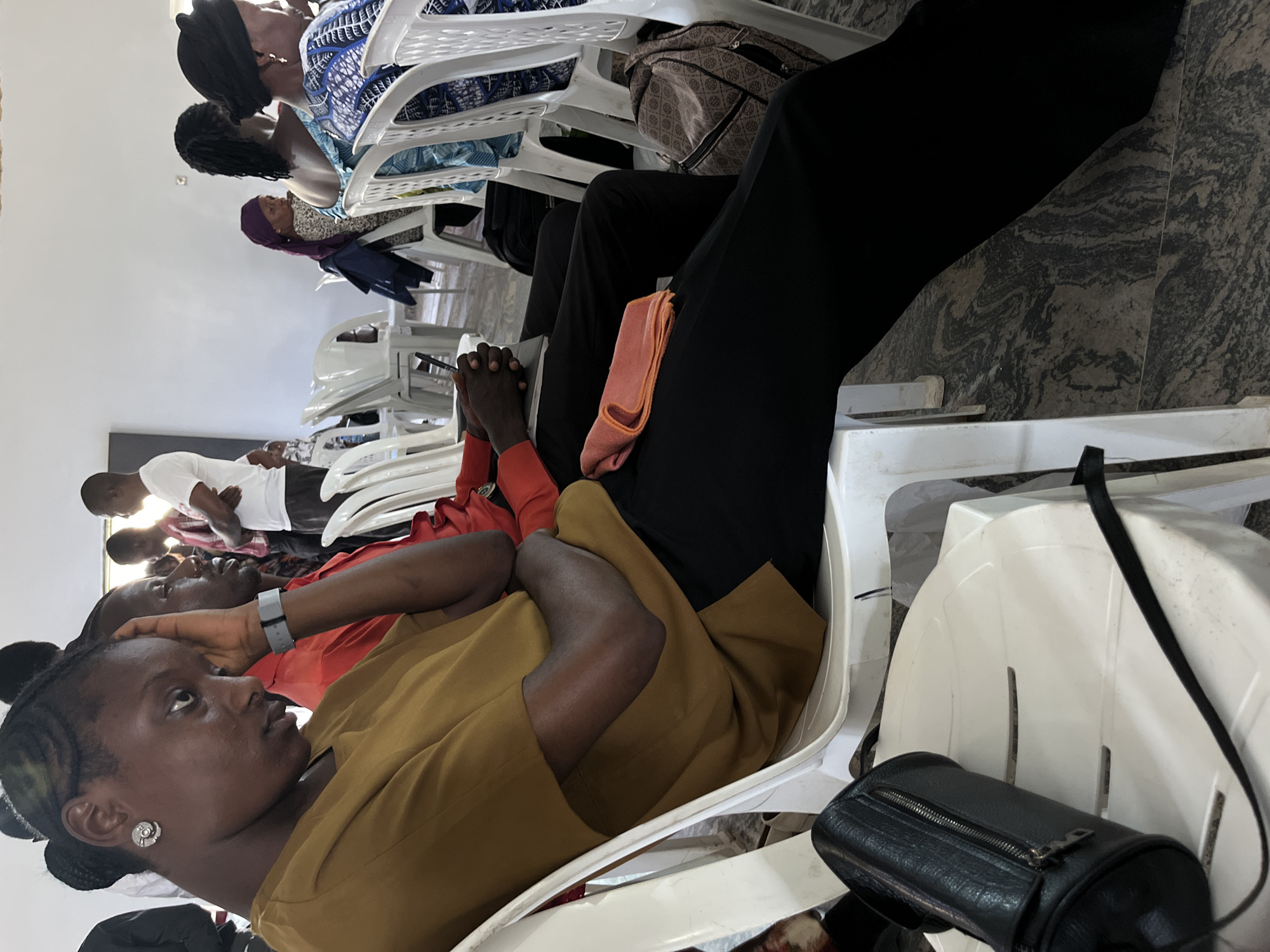 A medium shot of a group of people sitting in white plastic chairs at an indoor event, likely in Barakin Akawo, Nigeria.
The main subject, a young African woman, is seated in the foreground, wearing a brown blouse. Her gaze is directed upwards and to the left.
Behind her is a man in a red shirt with a wristwatch visible. Further back, additional people are seated, including individuals wearing blue patterns.
Objects present include a small black purse hanging on the side of one of the chairs, a beige handbag, and a peach-colored item placed on a black pant leg.
The setting appears to be an indoor space with off-white walls and a grey marble floor. The chairs are arranged in rows, suggesting a formal or semi-formal gathering.