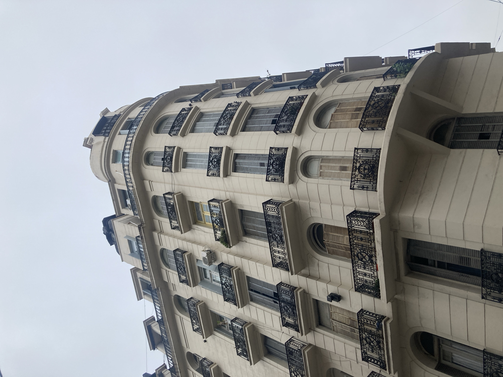 This low-angle, upward shot captures a multi-story building with a distinctive, smoothly curved facade, likely an apartment or residential block. The building is constructed from a light-colored material, possibly cream-colored stone or stucco, and features numerous windows with varied shapes. Some windows are rectangular, while others have an elongated oval or arched design.
A prominent detail is the elaborate black wrought ironwork, forming decorative balconies or railings in front of many windows. These railings exhibit intricate patterns and scrollwork. Many windows are fitted with closed shutters, some appearing to be louvered, others solid. A few windows show clear glass, and small air conditioning units are visible mounted below some windows on several floors. Sporadic greenery from potted plants can be seen on a few of the small balconies.
The sky above is a uniform light gray, indicating an overcast or cloudy day, with diffused light illuminating the building and casting no strong shadows. Faint overhead wires or cables are visible against the sky near the upper sections of the building. There are no people or discernible activities taking place. The architectural style, with its elegant curves and ornate ironwork, suggests an older urban environment. No visible text is present in the image.