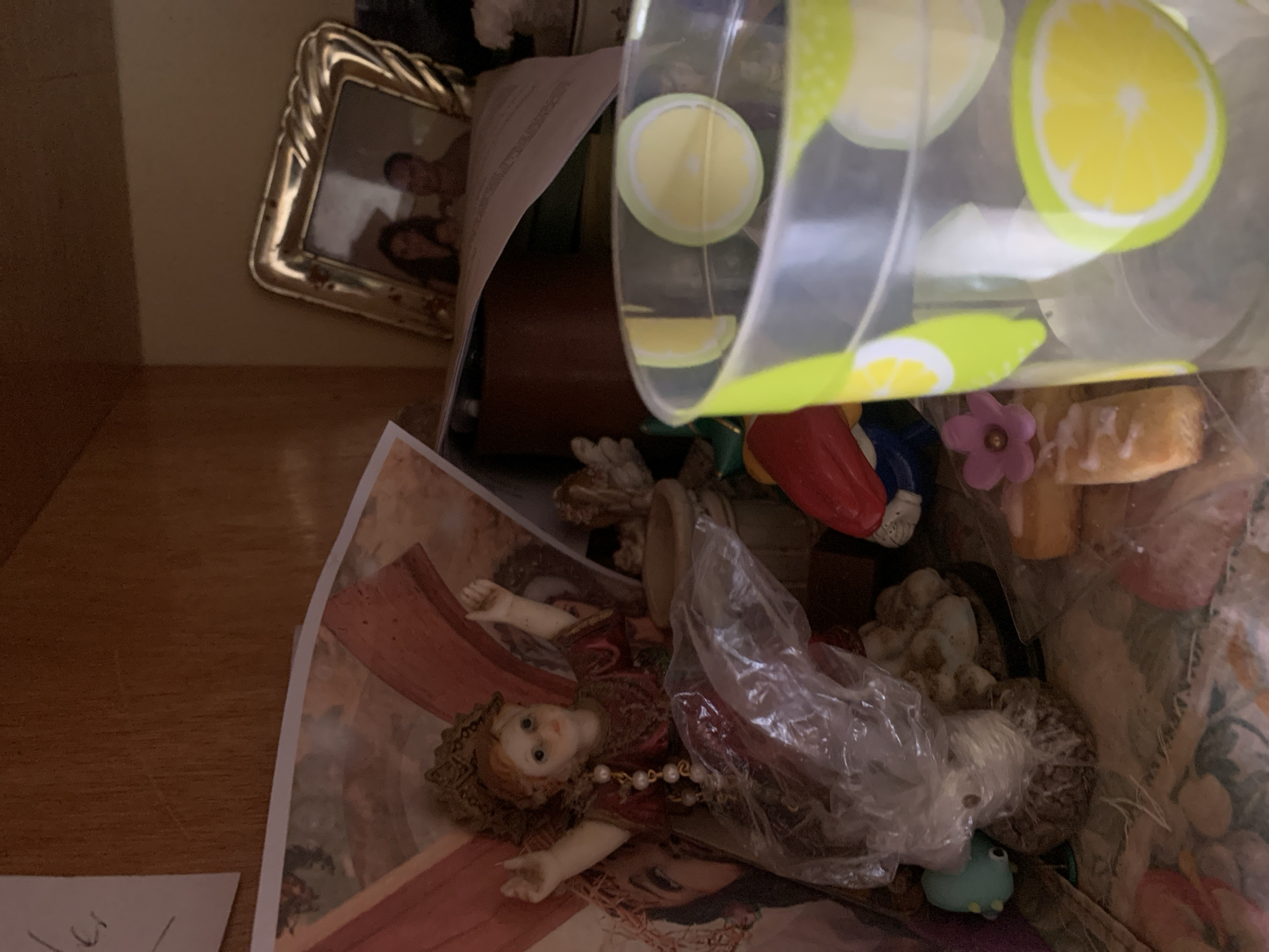 This close-up image, rotated approximately 90 degrees counter-clockwise, displays a cluttered collection of personal and decorative items, likely stored on a shelf, in a box, or a drawer. The setting is an indoor space in Santa Ana, Venezuela.
In the upper left quadrant, an ornate golden picture frame with a twisted design holds a photograph featuring two partially visible adult faces, one appearing to be a man and the other a woman. Behind this frame, a document with unreadable text is partially visible.
A clear plastic container, possibly a cup or small bucket, dominates the upper right and central area. It is decorated with bright yellow and lime green lemon slice patterns and seems to be holding or partially spilling other contents.
The central and lower parts of the image are filled with various figurines and decorations. A prominent porcelain or ceramic doll-like figurine, dressed in a maroon and gold outfit with a pearl necklace, lies on its back, exhibiting light skin, reddish-brown hair, and blue eyes. This doll rests on a larger printed image or poster, possibly depicting a classical artwork with warm reddish-brown hues.
Additional items include: a small, light blue/teal frog or toad figurine near the bottom; several white, textured objects that resemble decorative coral or stones; a red and blue decorative piece; a small purple flower decoration; and a white object with a textured, icing-like surface. A crumpled clear plastic bag is draped over some of the items.
No specific activity is taking place; the image captures a static arrangement of belongings. The lighting suggests an indoor environment. No significant text is clearly readable on any of the visible items or papers. The collection appears to consist of items with decorative or sentimental value.