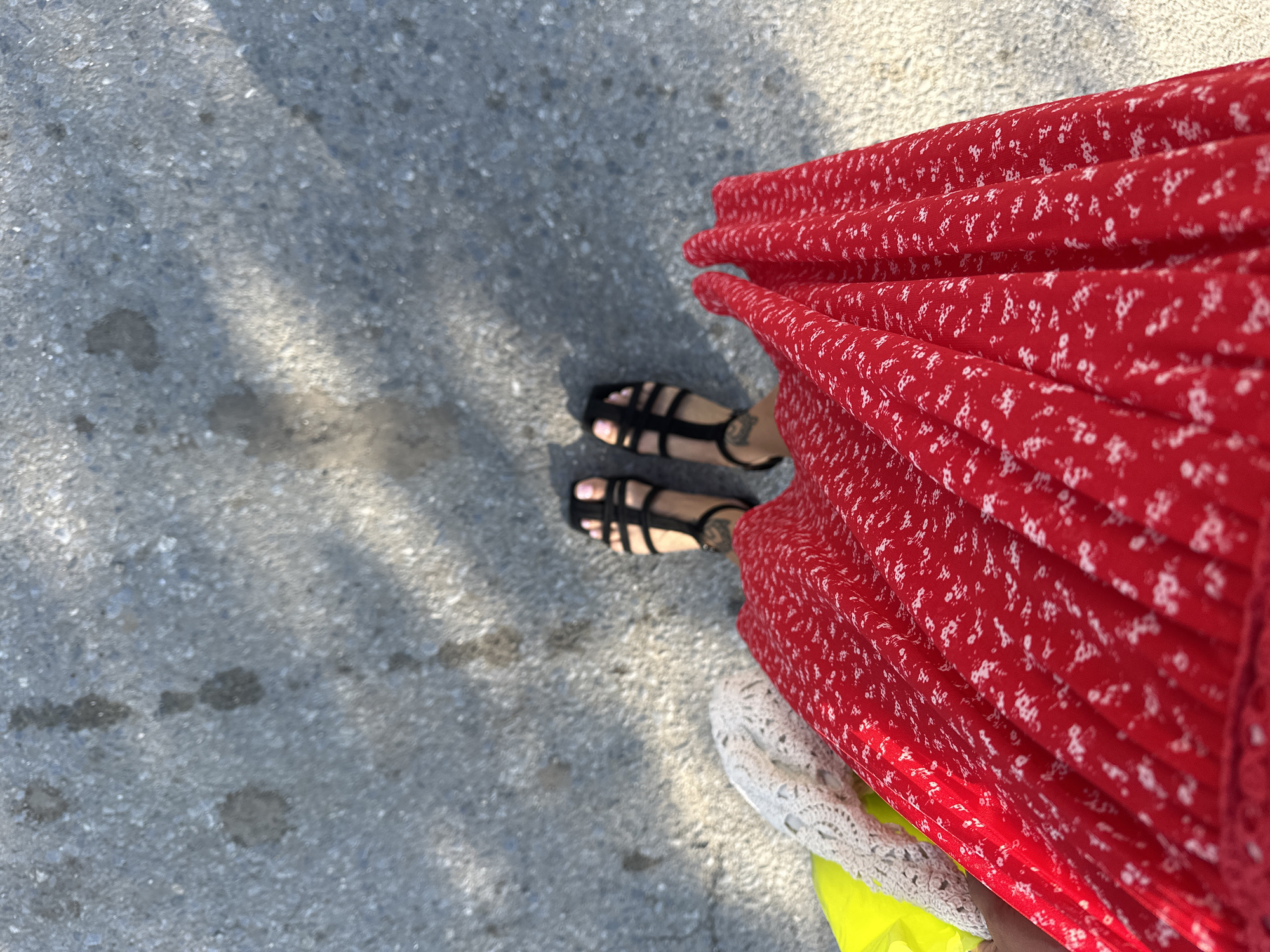The image shows a person's legs and feet standing on a gray asphalt surface. The person is wearing black strappy sandals and a red dress with a white floral pattern. A yellow bag and a white lace item are visible to the left of the dress. Shadows from foliage are cast on the asphalt. The scene appears to be outdoors. The location is Duvankul', Russia.