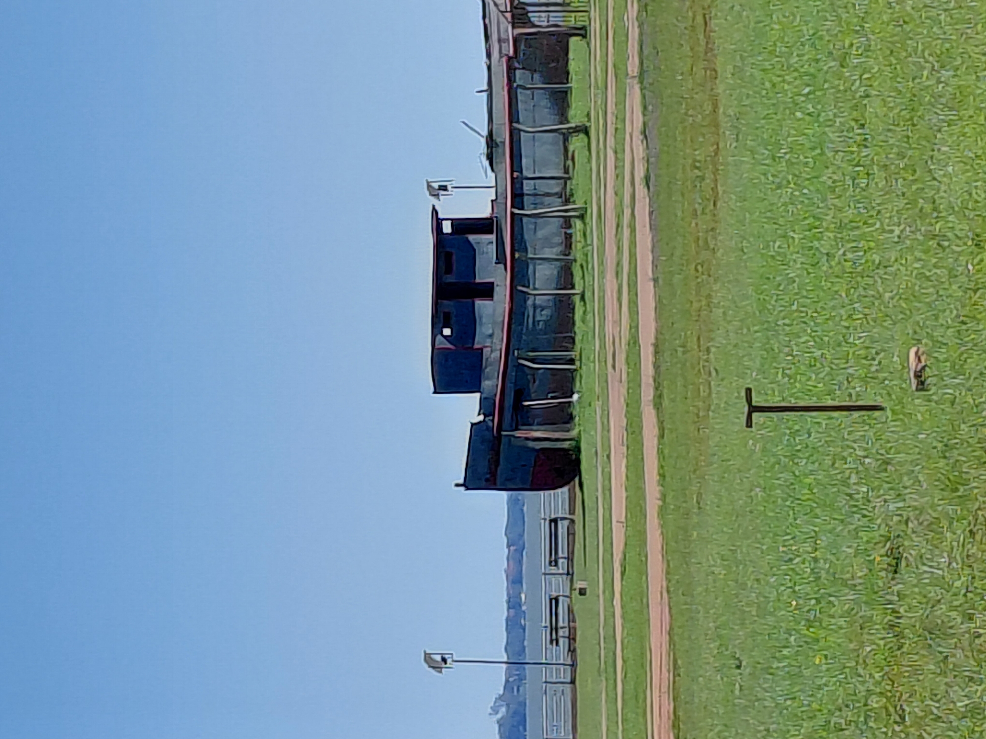 The image depicts a large, dark-colored, dilapidated boat structure situated on a grassy field under a clear blue sky. The boat appears to be partially enclosed by wooden posts and a fence-like structure. In the background, a body of water, possibly a river or lake, is visible, with a distant shoreline and some buildings. A single lamppost with a solar panel stands to the left of the boat. The foreground consists of well-maintained green grass with some dirt paths winding through it, and a T-shaped metal object is embedded in the grass. The overall scene suggests an outdoor area near the waterfront in Encarnación, Paraguay, likely a park or recreational space where the boat has been placed as an exhibit or landmark. There are no people or animals visible in the image.
